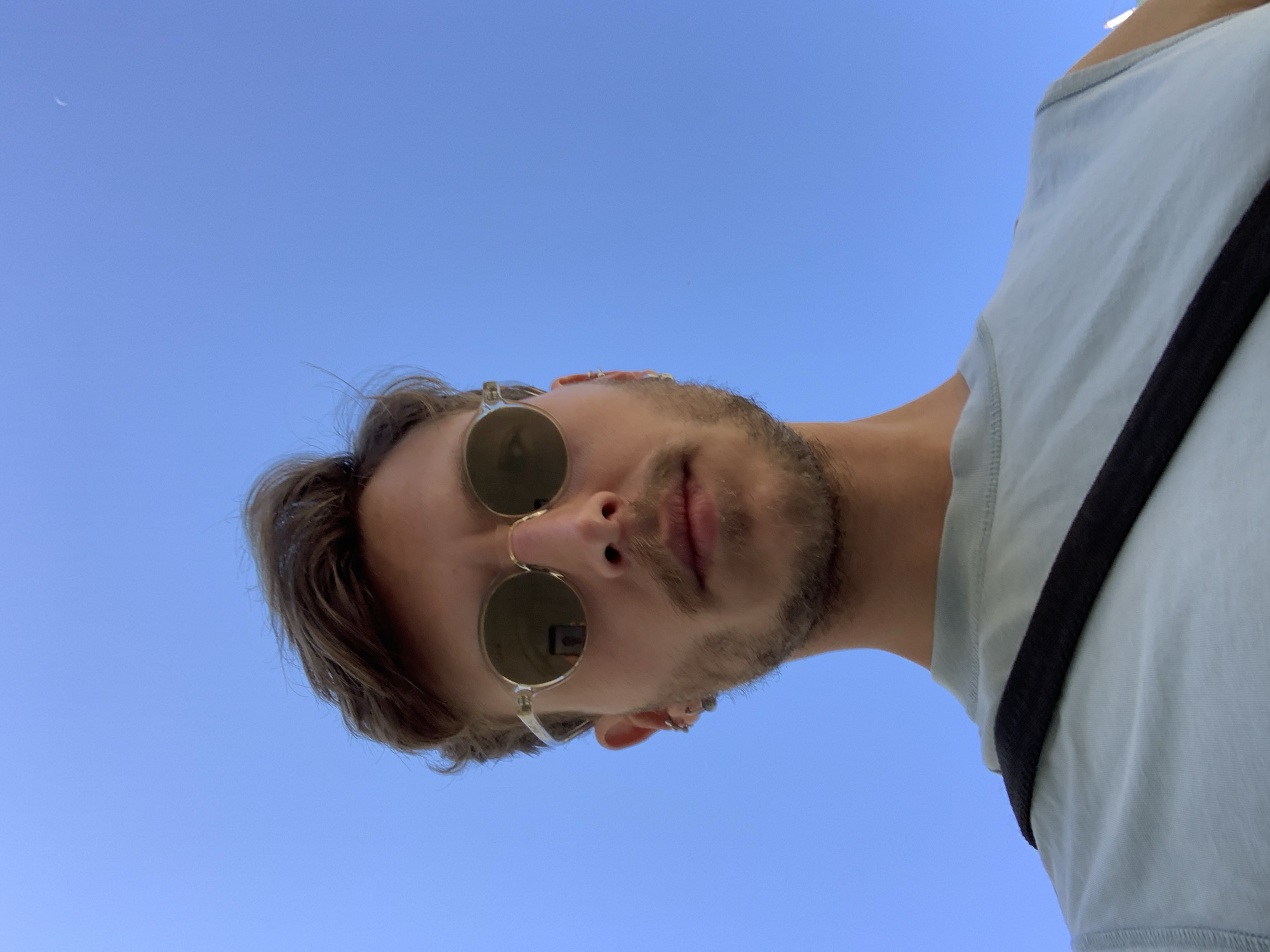 This image is an upward-angled selfie of a young man, presented rotated 90 degrees counter-clockwise. The subject, seen from below, has light brown hair, a short beard or stubble, and full lips. He is wearing round, gold-rimmed sunglasses that reflect a small object, likely a phone or camera, in the right lens. An earring or ear-cuff is visible on his left ear. He is dressed in a light blue or grey t-shirt, with a black strap, possibly from a bag or camera, extending across his right shoulder.
The background is a clear, vibrant blue sky, indicating daytime. A faint white crescent moon is visible in the upper left portion of the frame. The overall atmosphere is bright and casual. The provided context indicates the location is Tel-Aviv, Israel. There is no visible text in the image.