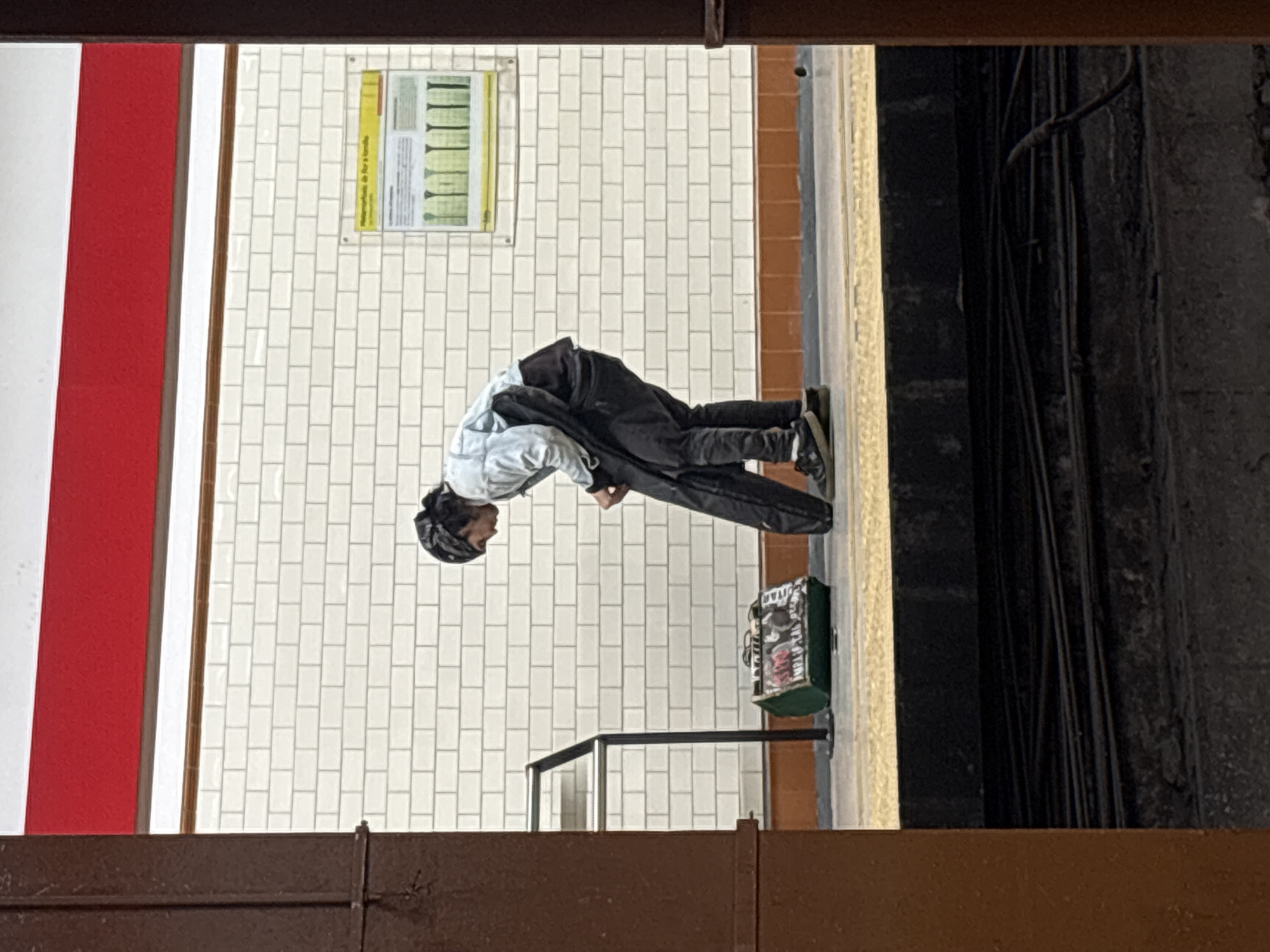 The image shows a person bending over on a subway platform, likely at a Buenos Aires, Argentina, subway station. The person is wearing a light-colored jacket, dark pants, and a hat. They appear to be picking something up. A green bag is on the platform next to them. The platform has white tiled walls with a poster and a red and white stripe. There is a metal railing nearby. The view is framed by a dark brown structure at the bottom and top of the image, as if taken from above or through a window. Wires are visible on a wall to the right of the platform.