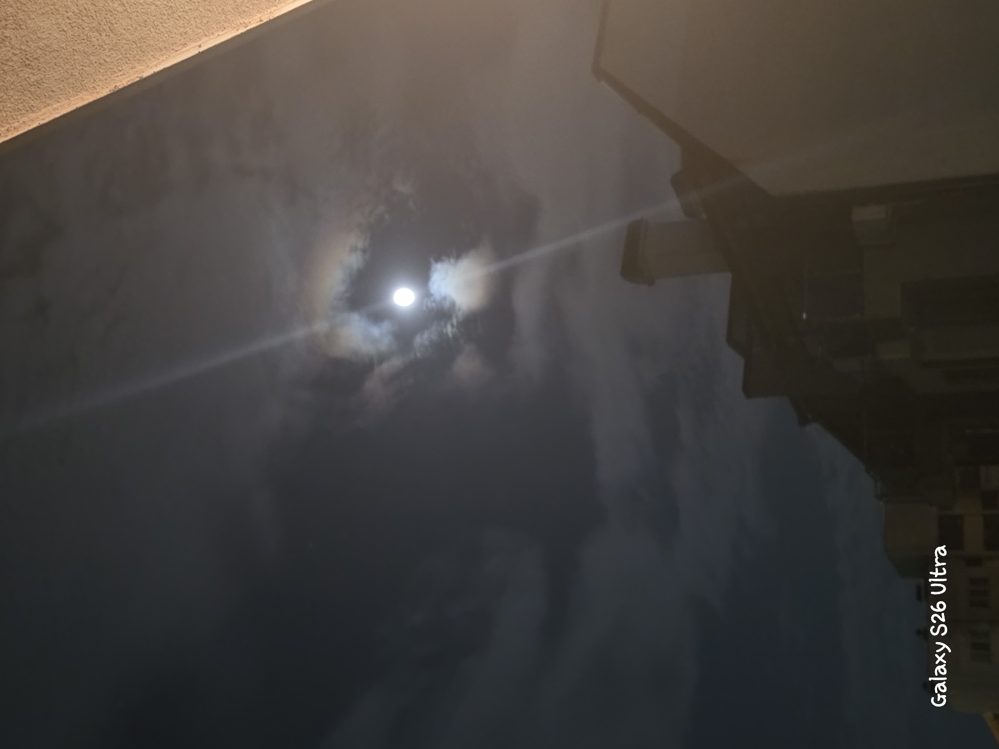 The image shows a nighttime scene with a bright full moon partially obscured by clouds. A light beam emanates from the moon, creating a halo effect. The moon is positioned in the upper left quadrant of the image, with a building's silhouette visible in the background. The building's reflection can be seen in the foreground, indicating a wet or reflective surface. The sky is dark, suggesting it is nighttime, and the presence of clouds implies overcast weather. The "Galaxy S26 Ultra" text is visible in the bottom right corner, oriented vertically.