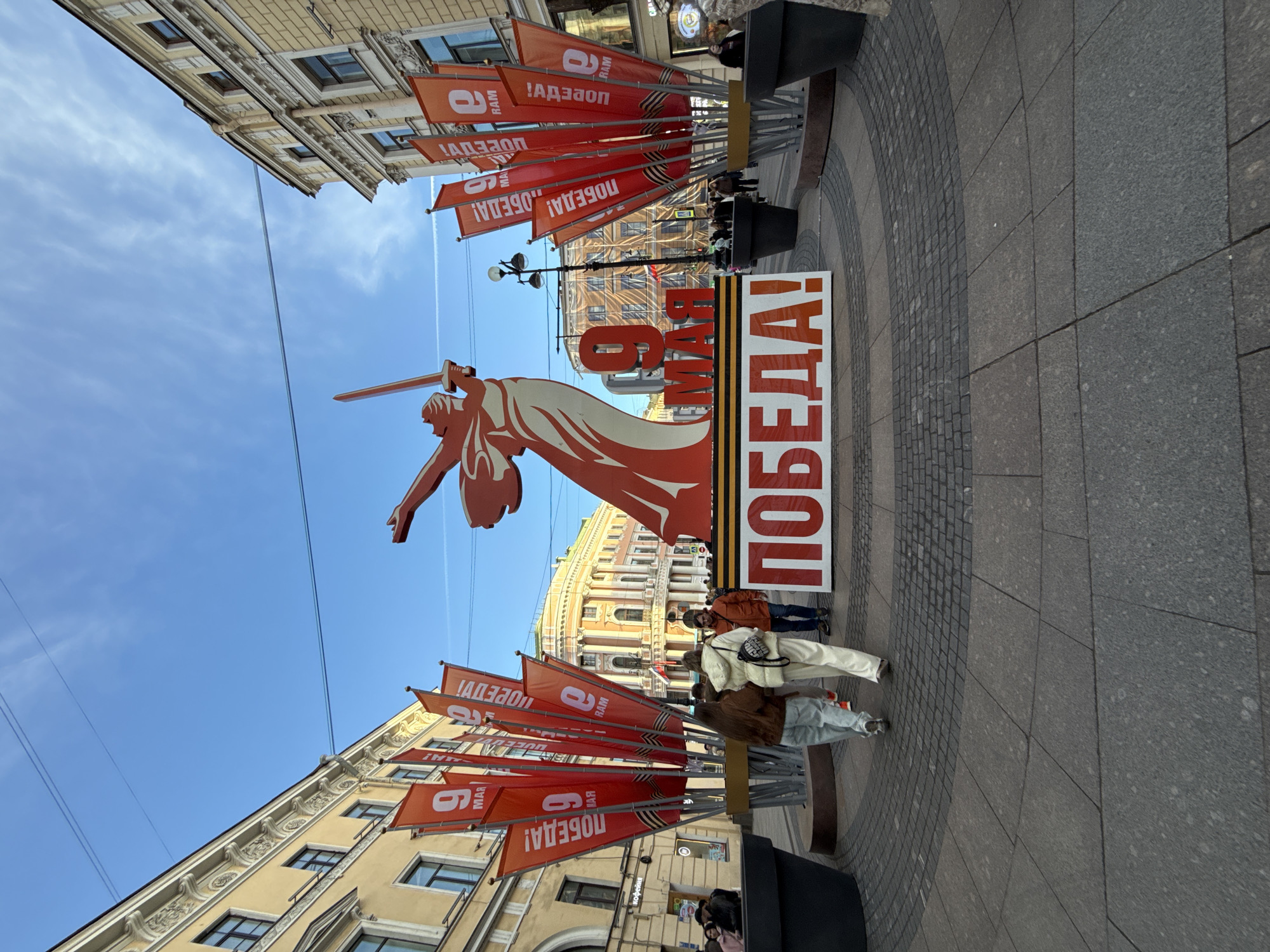 The image shows a street scene in Saint Petersburg, Russia, decorated to commemorate Victory Day on May 9th. A large, orange, stylized monument with the figure of "The Motherland Calls" statue holding a sword is featured, alongside a vertical sign that reads "May 9th Victory!" in Russian. Strings of red flags with the word "Victory!" in Russian, along with the date "9th May," are hung between buildings. Several people are walking along the sidewalk in front of the decorations. The buildings in the background suggest a typical urban streetscape with classical architecture. The street is paved with stone tiles in various shades of gray.