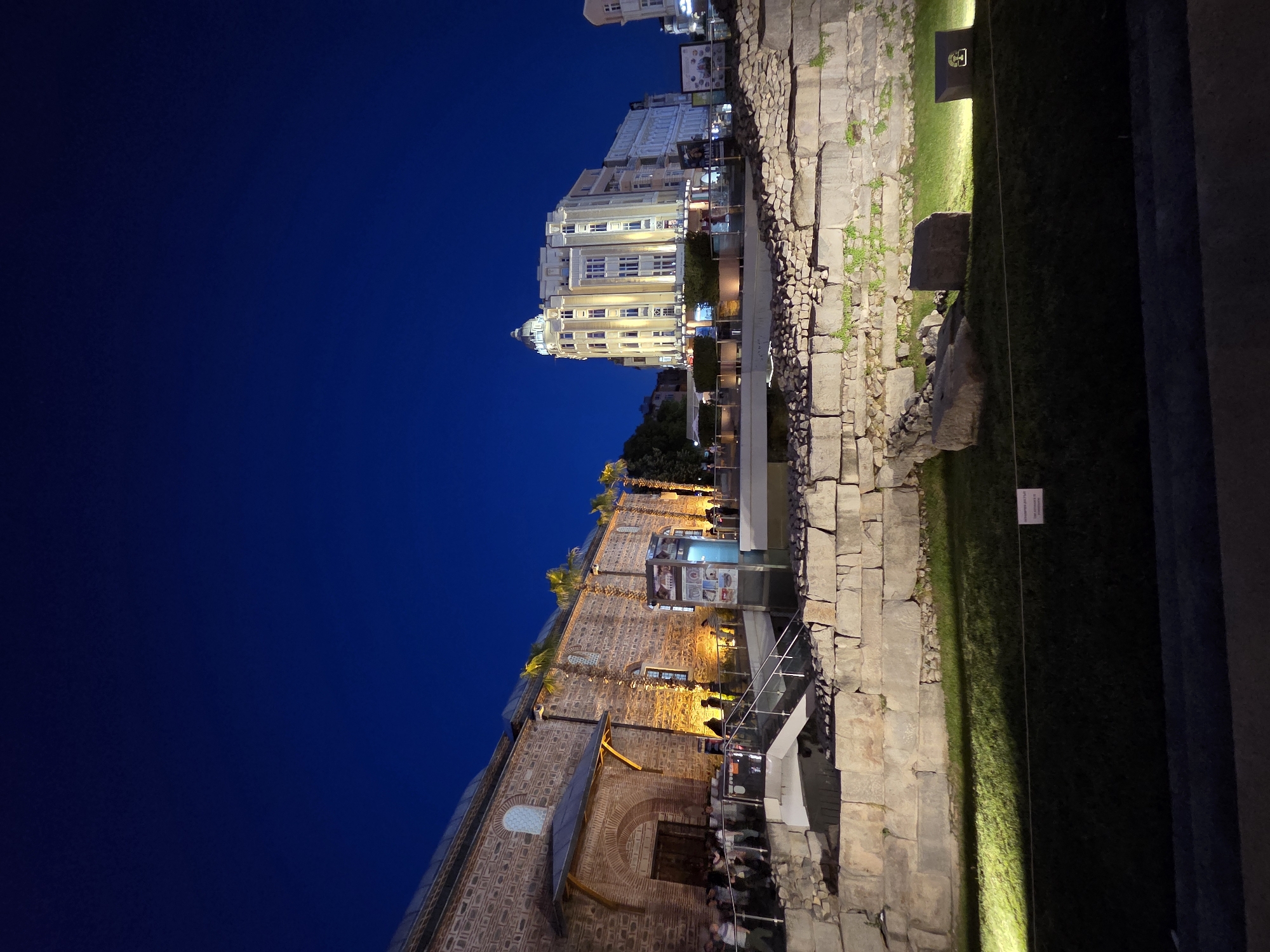 At night in Plovdiv, Bulgaria, a view shows the Eastern Gate of Philippopolis and the illuminated building of the Municipality of Plovdiv in the distance. People gather around the Eastern Gate, a historical site with exposed stone walls, adjacent to a modern structure with glass railings and informational displays. To the right of the gate is a grassy area illuminated by a spotlight, with segments of stone walls and informative plaques. The sky is a deep blue, indicating the darkness of the evening.