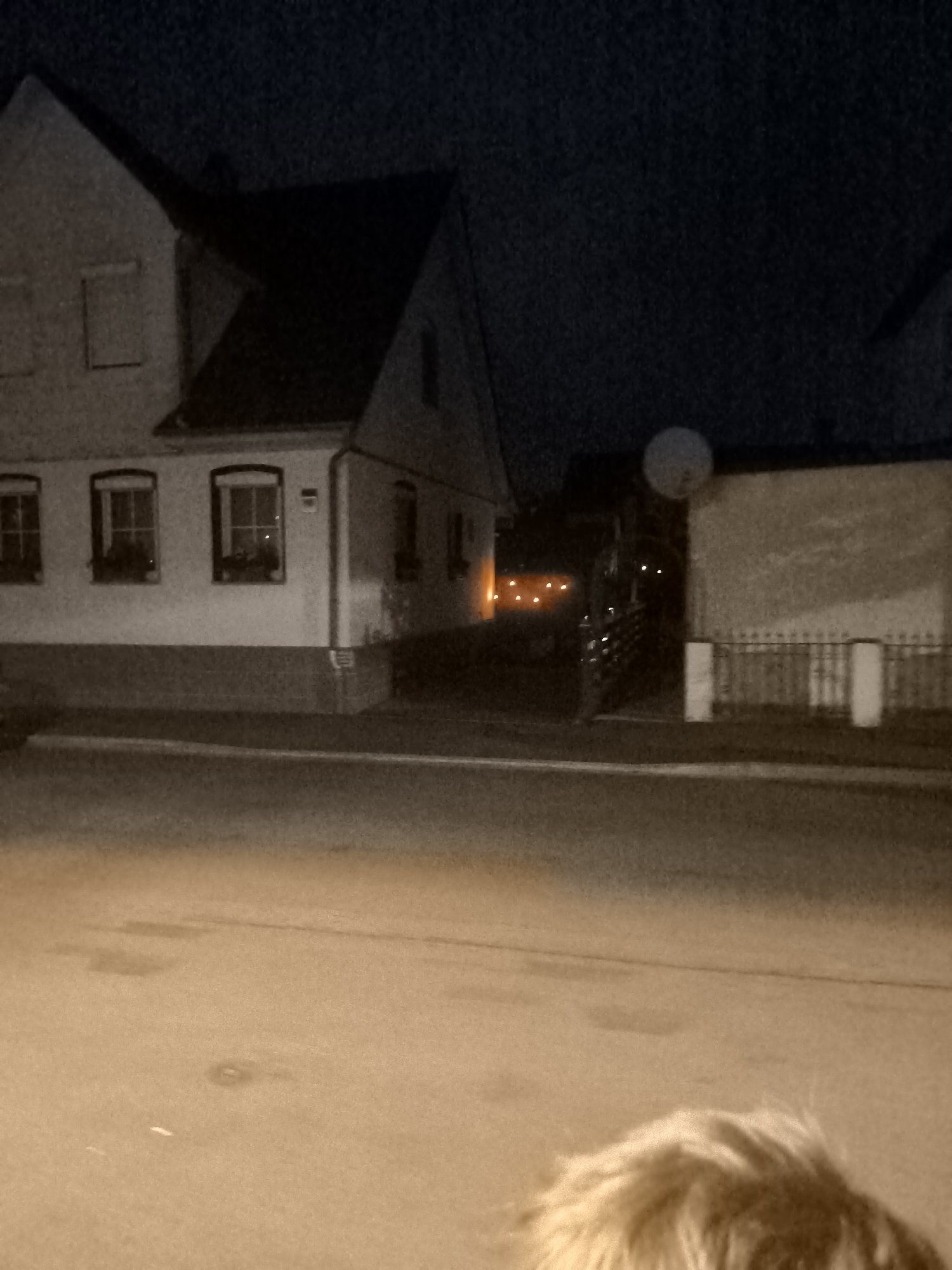 The image depicts an exterior scene at night in a residential area. In the foreground, the top of a person's blonde, possibly furry, hair is visible, suggesting the photo was taken from the perspective of someone looking over their shoulder or from behind them.
The middle ground shows a street with a sidewalk and a building that appears to be a house or a small apartment building. The building has multiple windows, some of which are illuminated from within with a warm, orange glow. The exterior walls of the building are light-colored, and there is a darker, brick-like foundation. Adjacent to the building, a fence with white posts and metal railings runs along the property line. Beyond the fence, a large, light-colored, dish-like object is mounted on the wall of another structure. Further in the background, more lights are visible, suggesting additional buildings or outdoor lighting. The sky is dark, indicative of nighttime. The overall impression is a quiet, possibly suburban or rural, nighttime setting.