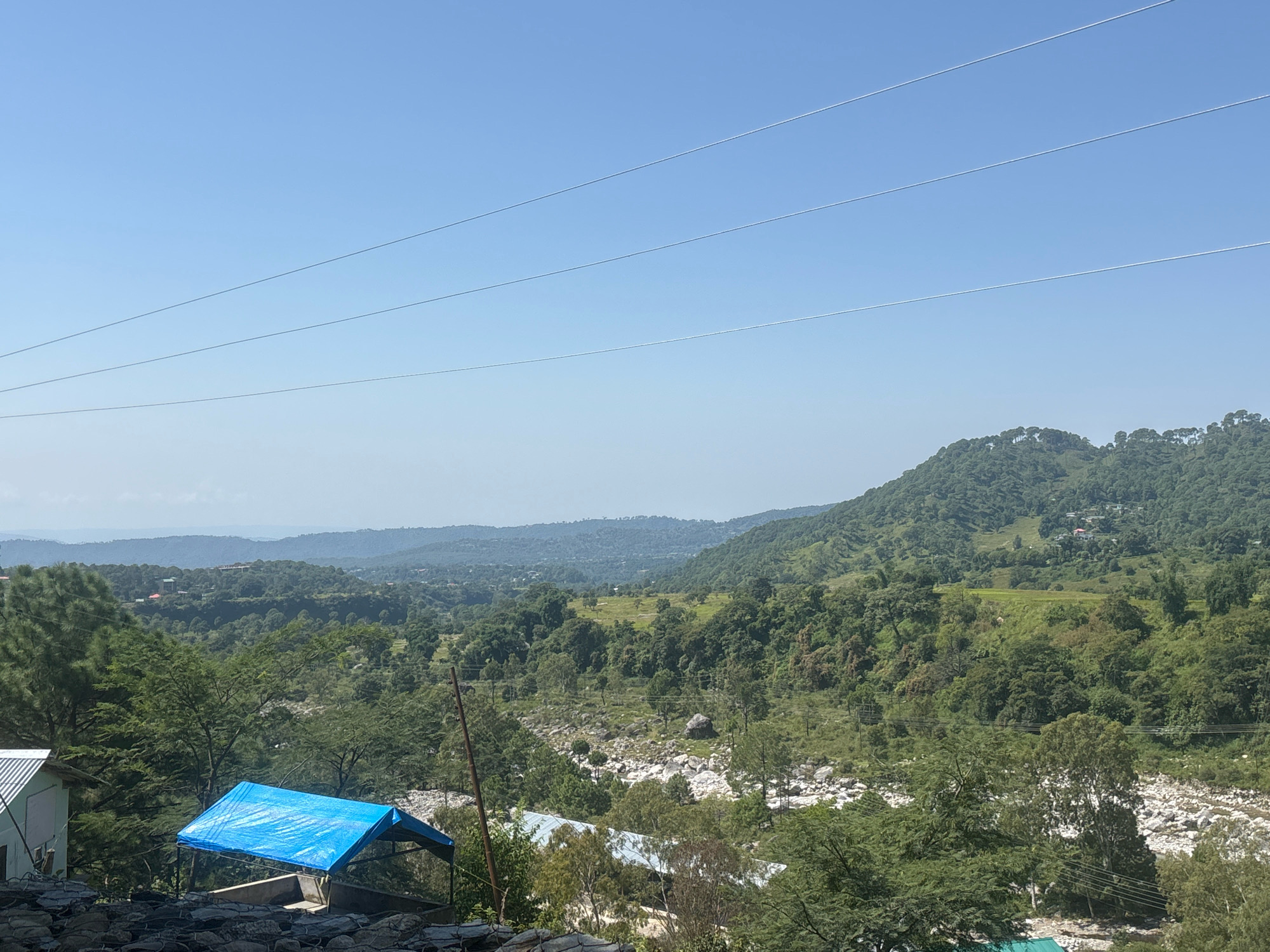 This is a high-angle, eye-level, medium shot of a lush, green valley in Palampur, India. The image is dominated by rolling hills covered in dense foliage and trees, with a clear blue sky overhead. In the foreground, a small section of a white building with a corrugated metal roof is visible on the left. Next to it, a structure covered by a bright blue tarp is erected. A dry, rocky riverbed cuts through the middle ground, and beyond it, the landscape continues to rise into more hills, dotted with scattered houses and agricultural fields. Three power lines stretch diagonally across the upper portion of the frame. The overall scene conveys a sense of rural tranquility and natural beauty.
