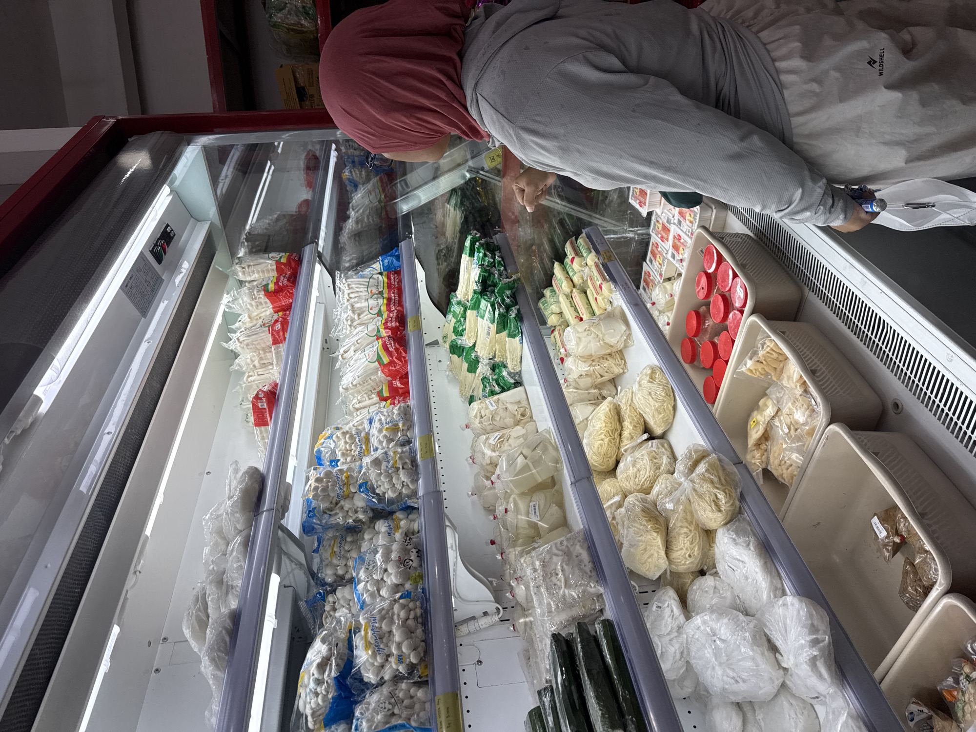 The image depicts a customer, a woman wearing a pink hijab and a grey hoodie, standing in front of a refrigerated display case in a supermarket. The display case is stocked with various packaged food items, including what appear to be vegetables, noodles, and possibly mushrooms. The items are neatly arranged on shelves and in bins. The woman is looking into the display case, with her right hand extended as if reaching for an item. She is holding a reusable shopping bag in her left hand. The supermarket appears to be well-lit, with bright overhead lighting. The location is identified as a supermarket in Tanjung Uban, Indonesia.