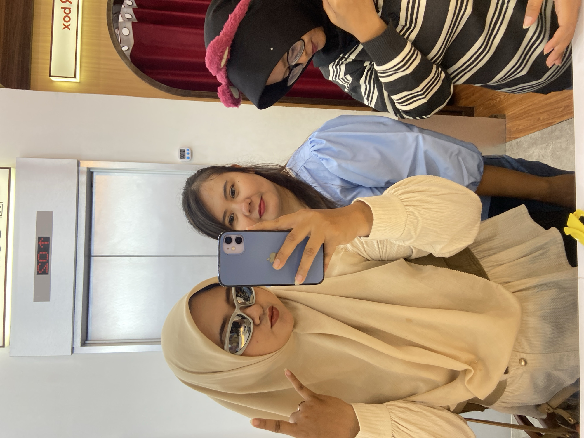 Three women are taking a selfie in front of an elevator in what appears to be a building, likely in Sidoarjo, Indonesia. The woman in the foreground wears a beige hijab, sunglasses, and a light-colored, textured long-sleeved top, gesturing with her left hand. The second woman, behind and slightly above the first, has black hair and wears a light blue blouse with puffy sleeves. The third woman, positioned at the top of the frame, wears a black beanie with pink trim, eyeglasses, and a striped black and white long-sleeved shirt. The woman in the middle holds a blue smartphone, capturing the selfie. The elevator's digital display reads "SOIT." The environment includes a hallway with a sign and decorative elements in the background.