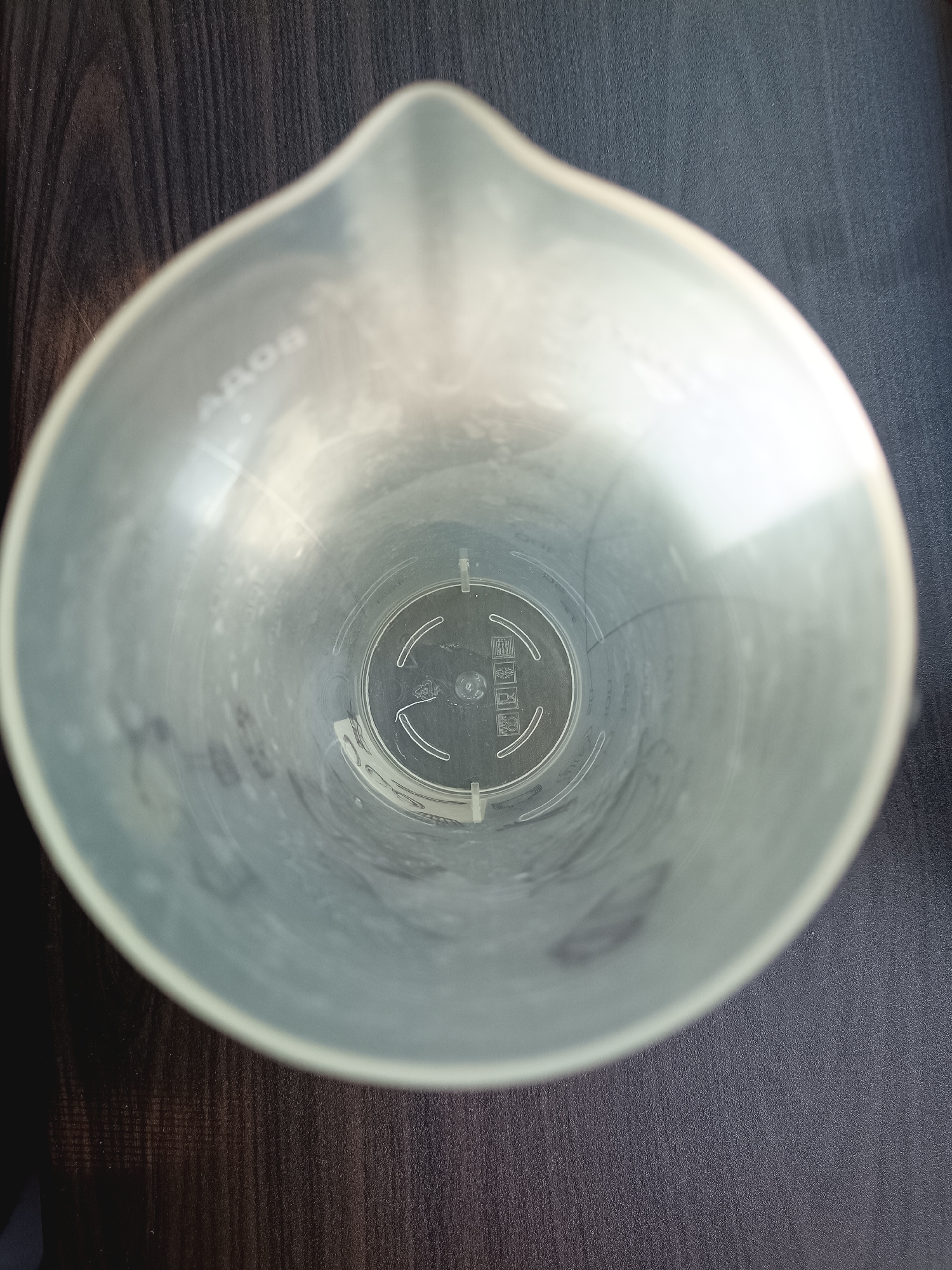 The image shows an overhead, close-up view of the inside of a clear plastic measuring cup. The measuring cup has a spout at the top. The interior walls of the cup are slightly opaque and show some smudges or residue. There are embossed markings on the inside bottom of the cup, including what appears to be a recycle symbol and some text that is difficult to read. The measuring cup is placed on a dark wood-grain surface, likely a table. The lighting is somewhat dim, casting shadows that suggest the light source is from the top right. The overall scene is static and there are no people or animals present. The image was taken in Donetsk, Ukraine.