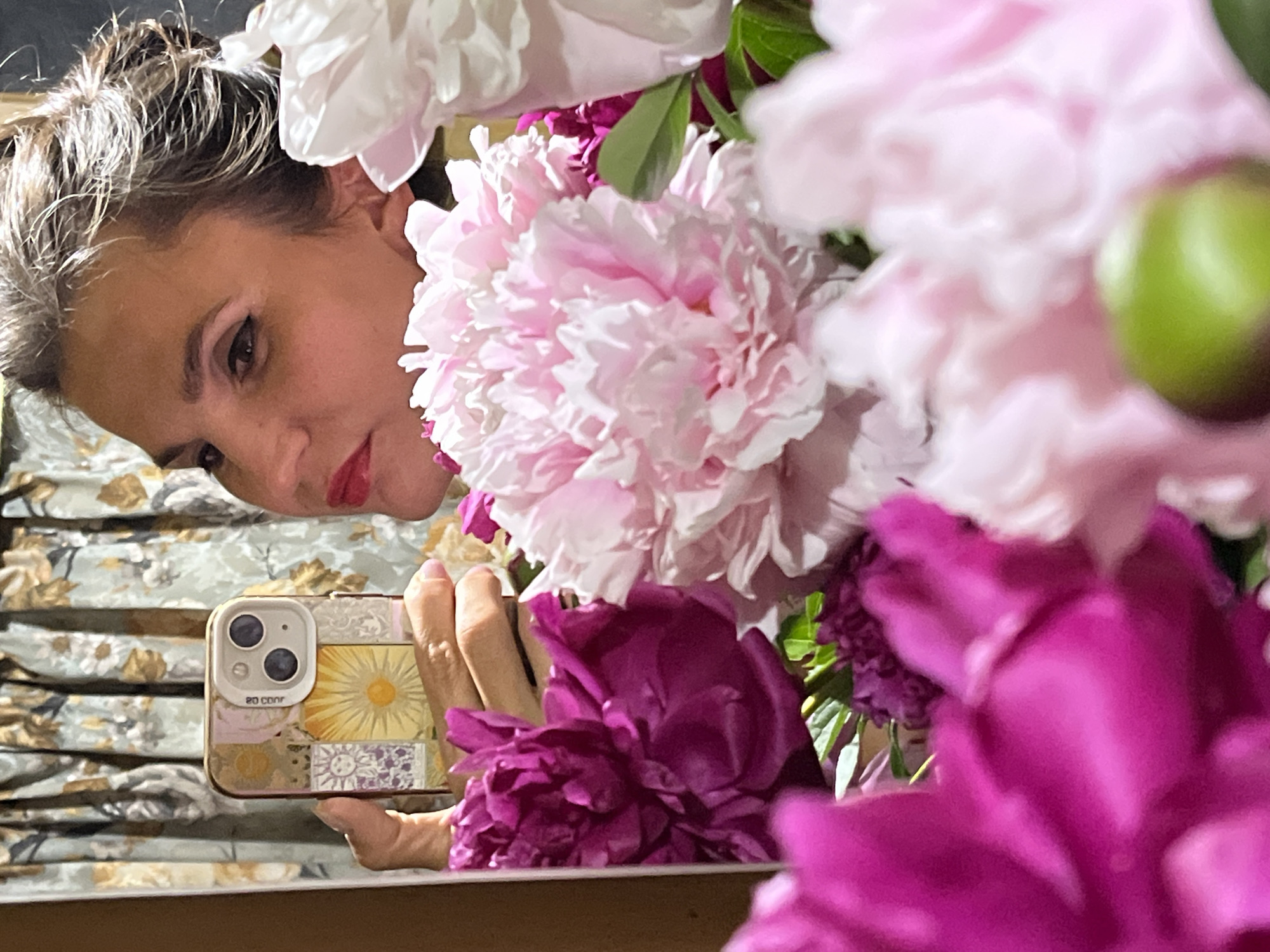 The image is a close-up, slightly overexposed selfie taken in Petropavlovsk-Kamchatsky, Russia. A woman, with her face visible in profile, holds an iPhone in her left hand, capturing her reflection in a mirror. She has dark hair pulled back, prominent eye makeup, and bright red lipstick. The phone case has a collage of sun-themed images. She is surrounded by a vibrant arrangement of peonies in various shades of pink and deep magenta. The background consists of a floral curtain. The overall impression is one of an intimate moment, possibly at home, with a focus on the woman and the lush flowers. The specific location within Petropavlovsk-Kamchatsky cannot be determined from the visual information provided.