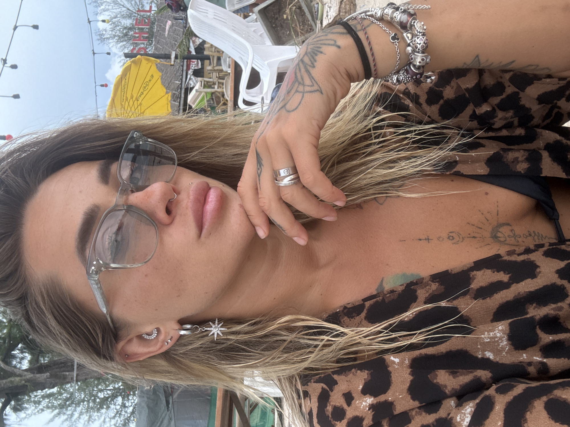 The image shows a close-up, head-and-shoulders shot of a woman with blonde hair and fair skin. She is wearing large, clear-framed glasses and has a nose ring. Her left ear has multiple piercings, including a crescent moon-shaped earring with rhinestones and a dangling star earring. She is wearing a leopard-print top over a black bikini top. Her right hand is resting on her chin, and she has several rings on her fingers, including a silver band. She has tattoos on her left arm and chest. The background appears to be an outdoor setting with trees, beach chairs, and a yellow umbrella. A sign that reads "SHELL" is visible in the distance. This scene is likely in Choeng Thale, Thailand, given the tropical vegetation and beach resort-like atmosphere.
