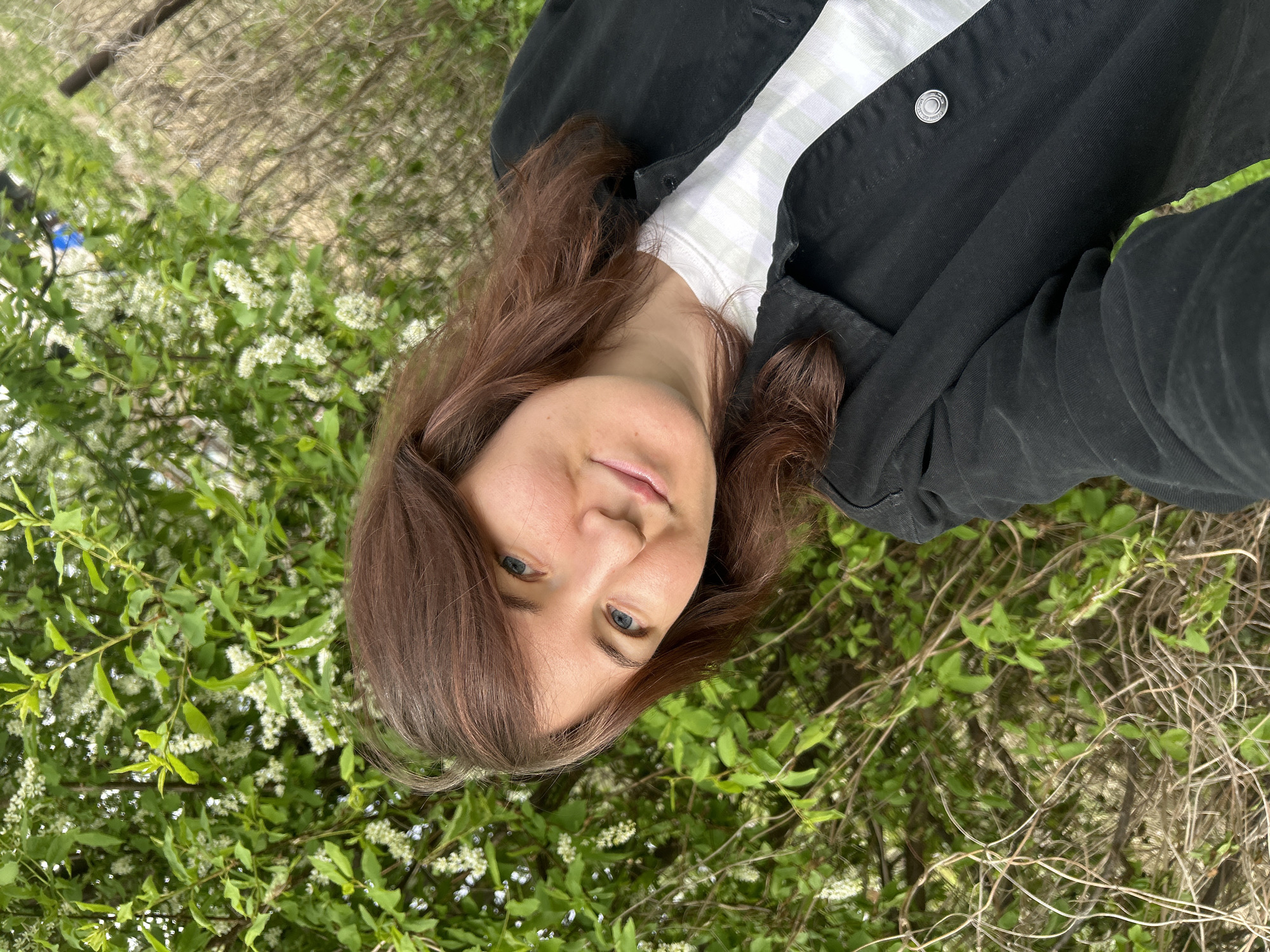 A woman with medium-length, auburn hair is centered in the image, angled slightly towards the viewer's left. She has fair skin and blue eyes, and is wearing a white top under a black jacket. The background consists of a bush with green leaves and small white flowers, some bare branches, and a hint of greenery. The photograph was not taken in Noviy, Russia.