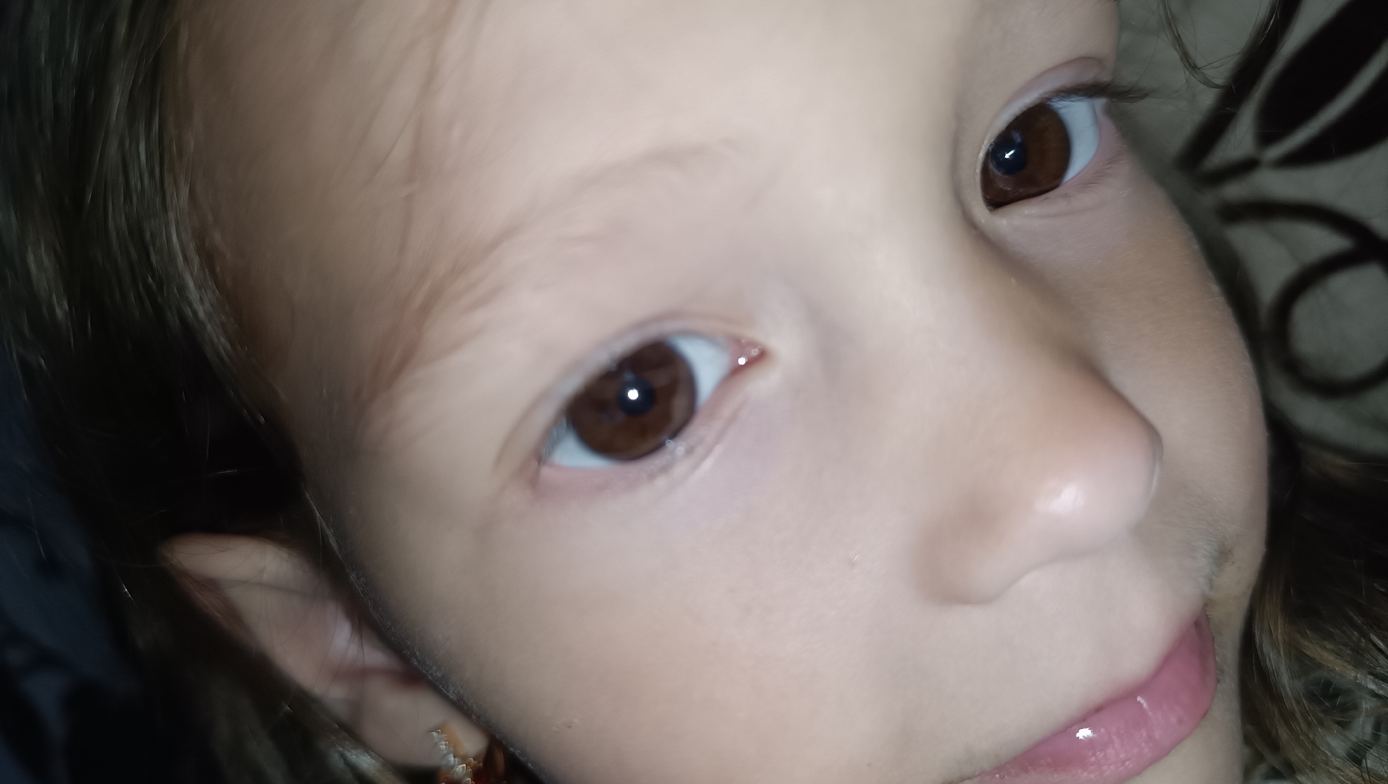 The image is a close-up of a young child's face, focusing on their eyes, nose, and mouth. The child has brown eyes and light skin. Their lips are slightly parted and appear to be glossy. The background is blurred and suggests a dimly lit indoor environment, with what looks like patterned fabric. There is no indication of the city of Karsha, Russia, in the image.