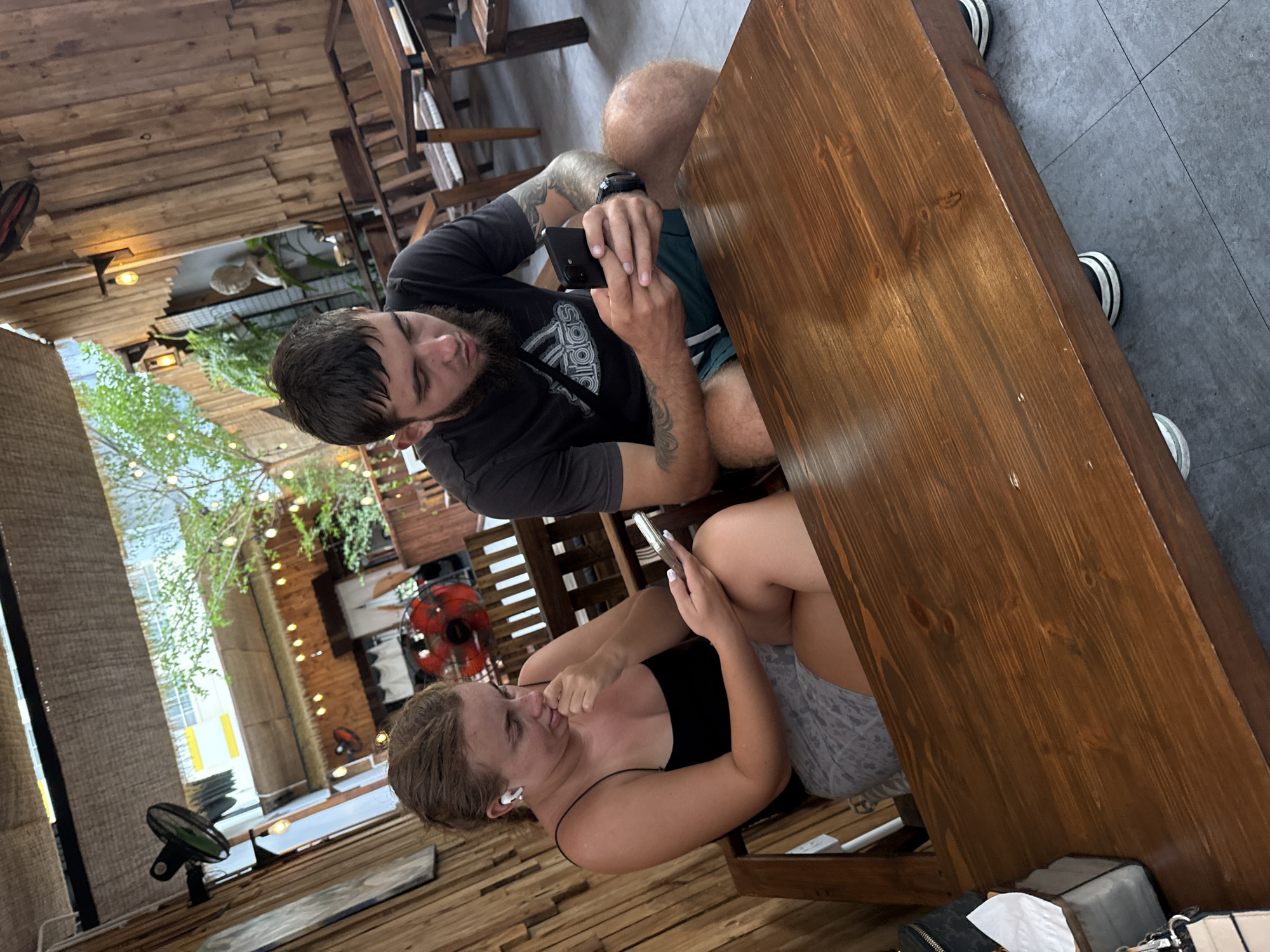Two individuals, a woman and a man, are seated at a wooden table in an outdoor cafe setting in Nha Trang, Vietnam. The woman, on the left, wears a black tank top and grey shorts, with white wireless earbuds in her ears. She is holding a smartphone in her right hand and appears to be interacting with it. The man, on the right, wears a dark t-shirt and green shorts and is also holding a smartphone, looking down at it. Both are focused on their devices, suggesting a moment of individual engagement. The cafe's architecture features rustic wooden walls and furniture, with a thatched roof overhead. Lighting from string lights and lamps is visible, creating a warm ambiance. In the background, a red fan is mounted on the wall, and lush greenery suggests a tropical environment. The overall scene captures a casual, everyday moment in a common urban cafe.