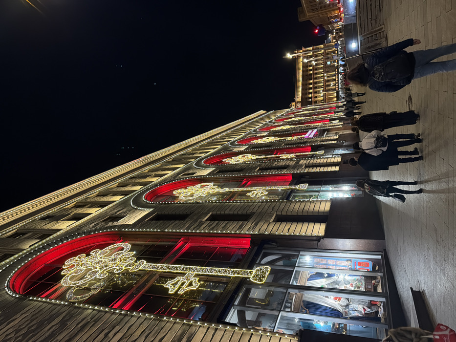 The image depicts a nighttime street view in Moscow, Russia, along the illuminated facade of the GUM department store adjacent to Red Square. The prominent building on the left is a multi-story structure with a textured, light-colored stone or masonry exterior. Its numerous large, arched windows are decorated with festive lighting: each arch is framed by warm white LED strings and features a red internal illumination. Within these red-lit arches, elaborate, vertically oriented key-shaped light installations, also composed of warm white LEDs, are suspended. The building's upper edges are similarly outlined with warm white LED lights. At ground level, a retail display window is visible on the far left, showing blue drapes and illuminated merchandise.
A wide, light-colored paved pedestrian walkway occupies the foreground and mid-ground. Approximately ten individuals are present on this walkway. In the immediate foreground, a person wearing a dark jacket and a backpack is seen from behind, angled slightly towards the left. Further into the mid-ground, a group of five individuals, dressed in dark cool-weather attire, walk away from the camera towards the right. Other pedestrians are visible as smaller figures further down the street. In the background to the right, another large, multi-story architectural building is distinctly lit with warm white lights. The sky is dark and clear, indicative of nighttime. Distant vehicle or streetlights are faintly visible on the far right.