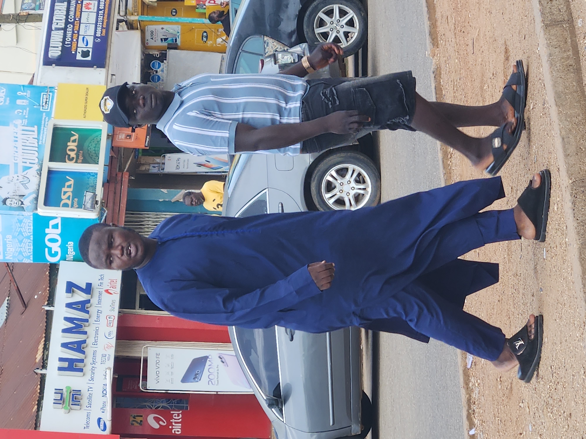 In Jos, Nigeria, two men walk on a dusty roadside. The man on the left wears a long, dark blue traditional outfit and black slides with a white "LV" logo. The man on the right wears a light blue and white striped short-sleeved shirt, black distressed denim shorts, and black sandals. Behind them, several businesses are visible, including "HAMAZ," which sells electronics and telecommunications services, and a yellow building displaying "OLUWA GLOBAL." Cars are parked and driving on the road. The scene appears to be a bustling commercial area.