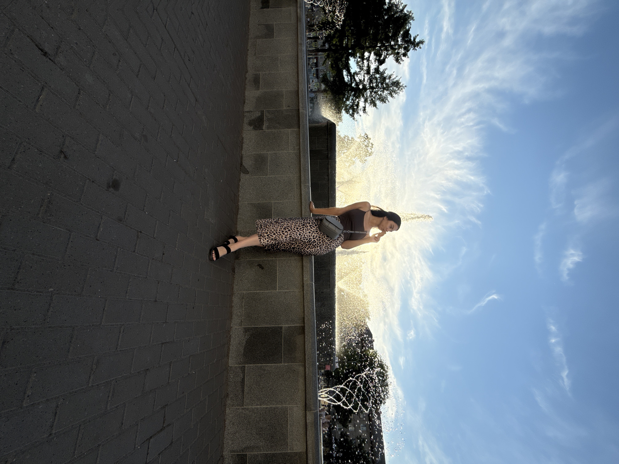 An outdoor full shot captures a woman sitting casually on a low grey stone wall next to an active fountain, under a bright blue sky with wispy white clouds. The scene is set in a public space, likely a park or square, in Chaikovsky, Russia.
The woman has dark hair and is dressed in a dark brown tank top, a leopard print midi skirt, and black sandals. A dark grey shoulder bag is slung across her body. She sits with her body slightly turned, looking downwards and to her right, with one hand gently touching her chin or mouth, giving a thoughtful or contemplative impression.
To her right, an elaborate fountain is vigorously spraying water upwards and outwards, creating a shimmering effect as the bright sunlight catches the droplets. The sun appears to be low in the sky, possibly late afternoon or early morning, backlighting the fountain and highlighting the water. Behind the fountain and slightly to the left, a dark green tree with visible branches adds depth to the background. The area beside the wall where the woman is seated is paved with dark grey brick-patterned blocks. The clear sky with scattered clouds suggests sunny and pleasant weather. There is no visible text in the image.
