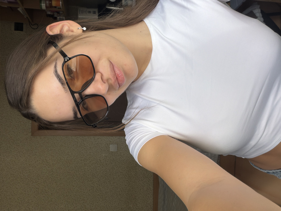 A female subject is captured in a medium-close-up, self-portrait photograph within an indoor residential setting. She has long, dark brown hair and fair skin, and is wearing black-framed aviator-style sunglasses with amber-tinted lenses, obscuring her eyes. Her lips are slightly parted, forming a subtle smile. She is dressed in a tight-fitting, white short-sleeved crop top, with a portion of light-colored denim fabric visible at the bottom left. A small, silver-colored stud earring is present in her right earlobe. The background to the left features a wall covered in light-colored, patterned wallpaper, containing a white electrical outlet and a partial dark wooden door frame. The background to the right is less distinct, showing elements suggestive of shelving or a cabinet unit with various items. Natural light illuminates the scene, casting shadows. The specific location within Sofia, Bulgaria, cannot be verified from the visual information provided in the image.