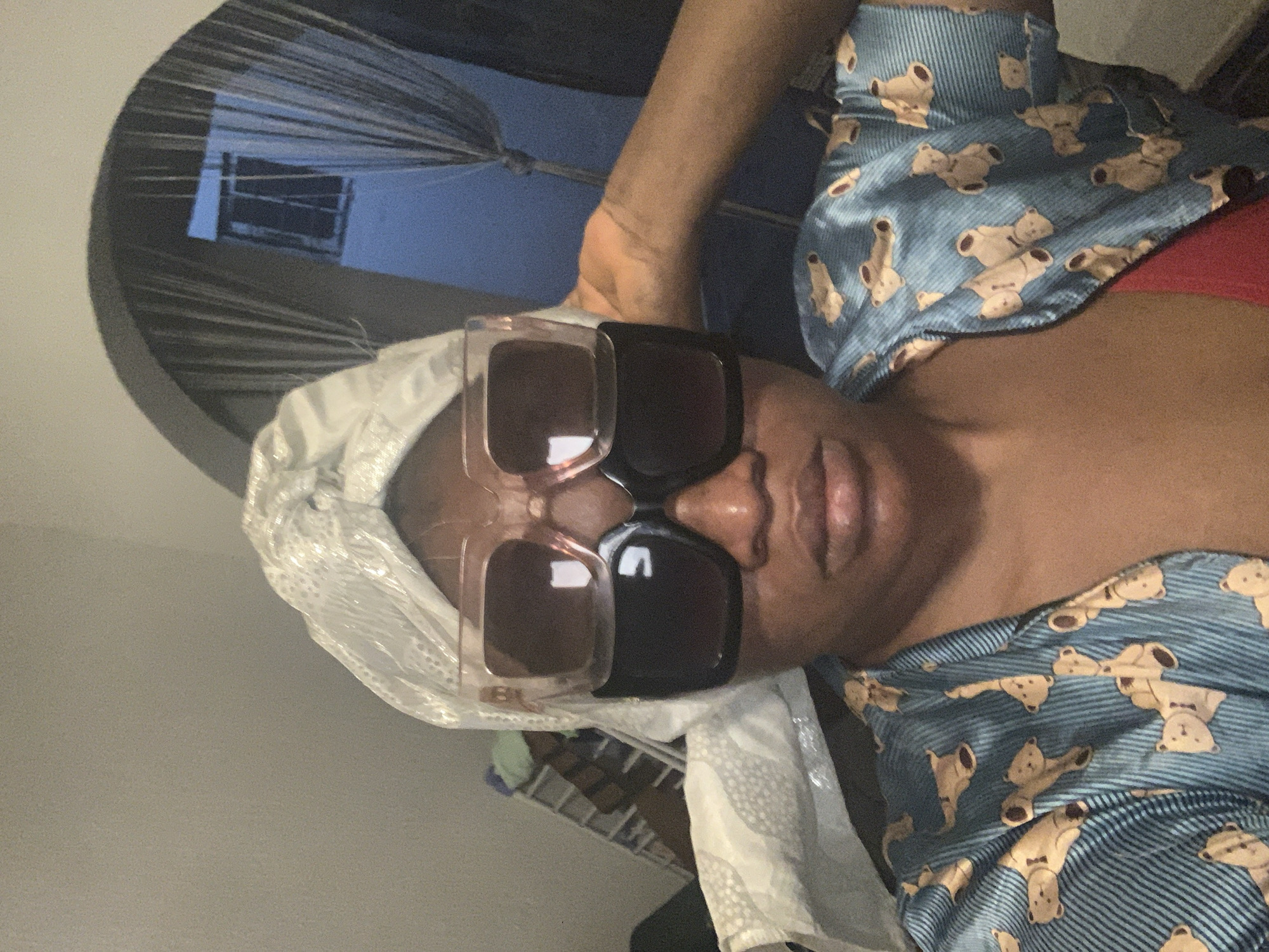 A person wearing sunglasses and a head covering is shown in a medium shot. The person is wearing a blue shirt patterned with teddy bears. The scene appears to be indoors. It is not possible to determine the location in Barakin Akawo, Nigeria from the given image.