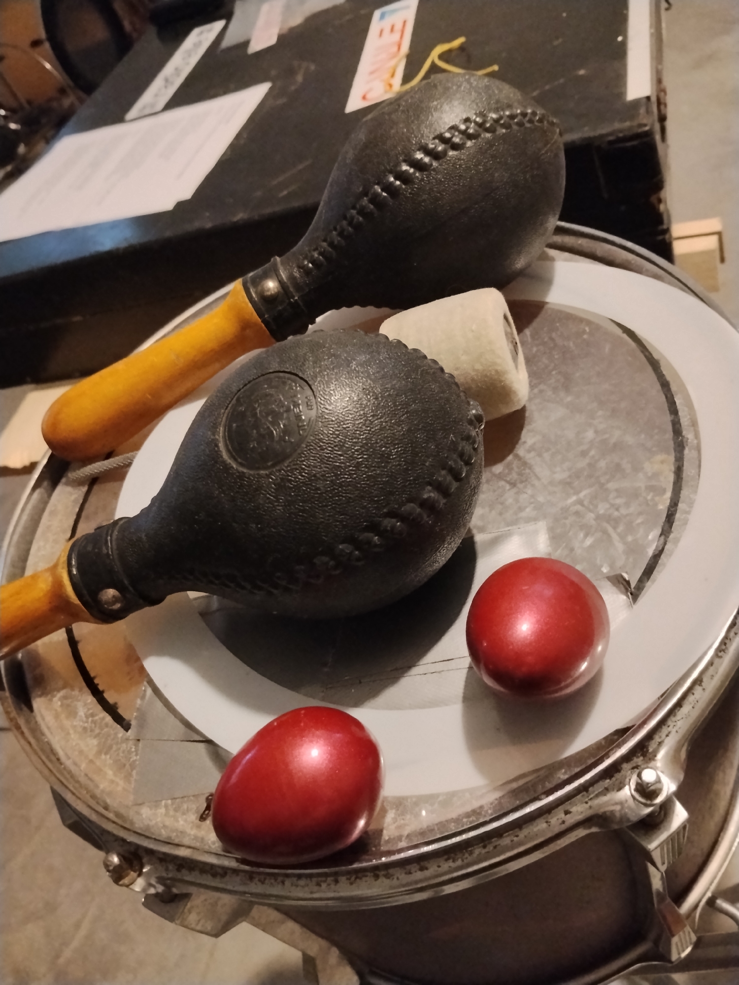 The image displays a close-up, overhead view of musical instruments resting on a drum. The primary objects are two maracas, one above the other, both with black, textured heads and wooden handles. The upper maraca is positioned diagonally, with its handle pointing towards the left. The lower maraca is oriented more horizontally, with its handle extending to the left and slightly downwards. Between the two maracas, on the drumhead, lies a small, cylindrical object with a light-colored, fuzzy texture, possibly a felt damper or a drumstick tip. To the right of the lower maraca are two small, shiny, red, egg-shaped objects, possibly decorative ornaments or small percussion instruments.
The instruments are placed on top of a drum, which has a reflective, light-colored drumhead and a silver rim with visible hardware. The drum appears to be set on a floor or surface with a textured, grayish appearance. In the background, a dark, flat surface is visible, possibly a case or a table, with some papers and labels on it. One label appears to have red and yellow markings, and another has printed text in what looks like English.
The lighting suggests an indoor setting, likely a practice space or storage area, with soft shadows indicating a diffuse light source. There is no indication of time of day or weather. The overall mood is static and focused on the arrangement of the objects. The location context of Santeramo in Colle, Italy, is provided but not visually evident within the frame. No people are visible in the image.