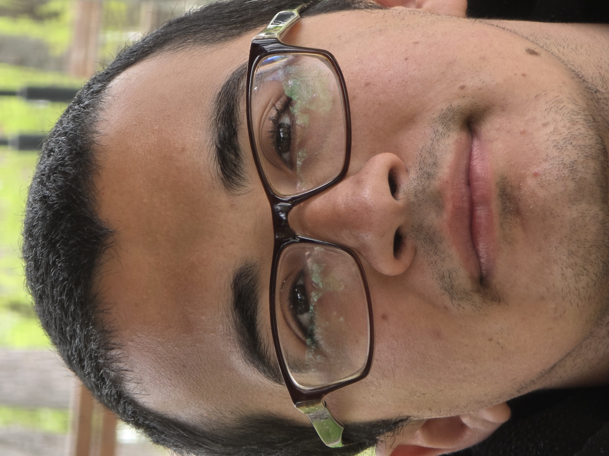 A close-up portrait shows a man with dark hair, dark eyebrows, and light skin wearing glasses with brown frames. The glasses reflect a green and light-colored background, suggesting an outdoor environment, potentially with trees or foliage. He has a slight stubble beard and his lips are closed in a subtle smile. The background is out of focus, further emphasizing the subject. The location is Unknown, Unknown.