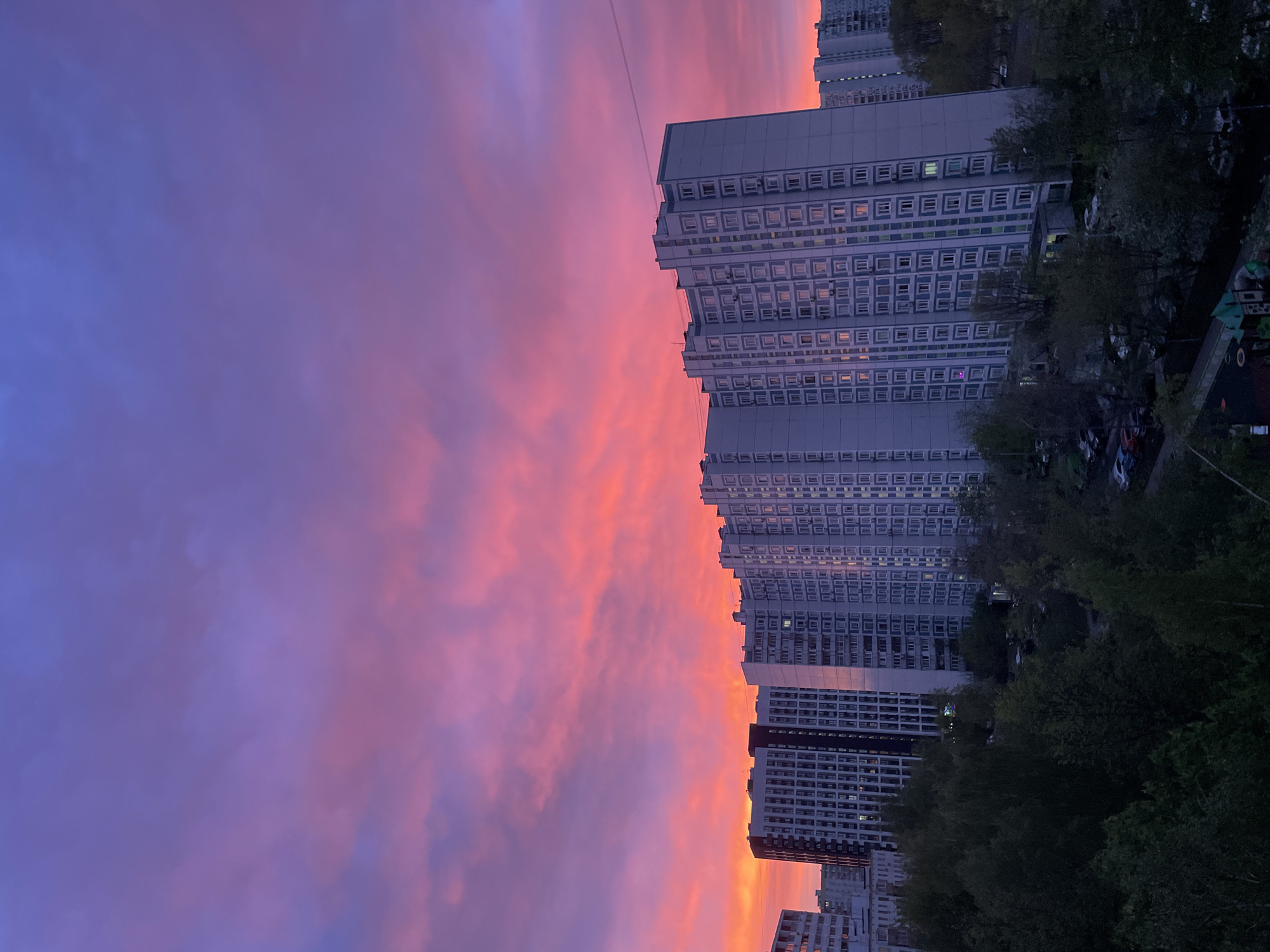 The image shows a cityscape with tall apartment buildings in Moscow, Russia, under a vibrant sunset sky with shades of orange, pink, and purple. The buildings have a repetitive, grid-like pattern of windows. Trees are visible on the right side of the frame.