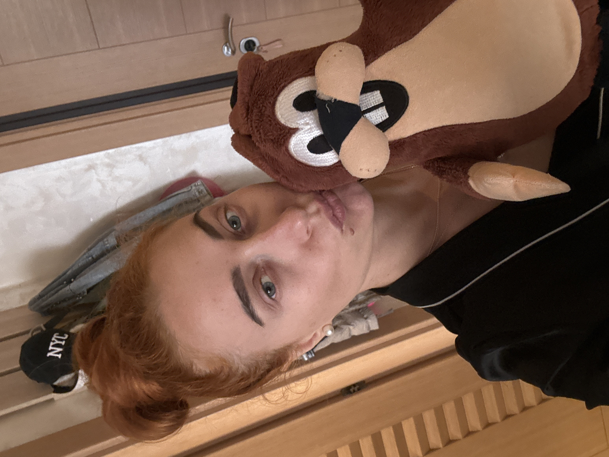 A woman with auburn hair and green eyes is holding a plush toy. The toy is a brown and tan cartoon animal with buck teeth. The woman is wearing a black shirt. A black baseball cap with "NYC" embroidered on it and a denim jacket are visible in the background. The setting appears to be indoors, near a wooden door or wall, possibly in a residential location, but no specific evidence places it in Budva, Montenegro. The interaction is holding the toy.