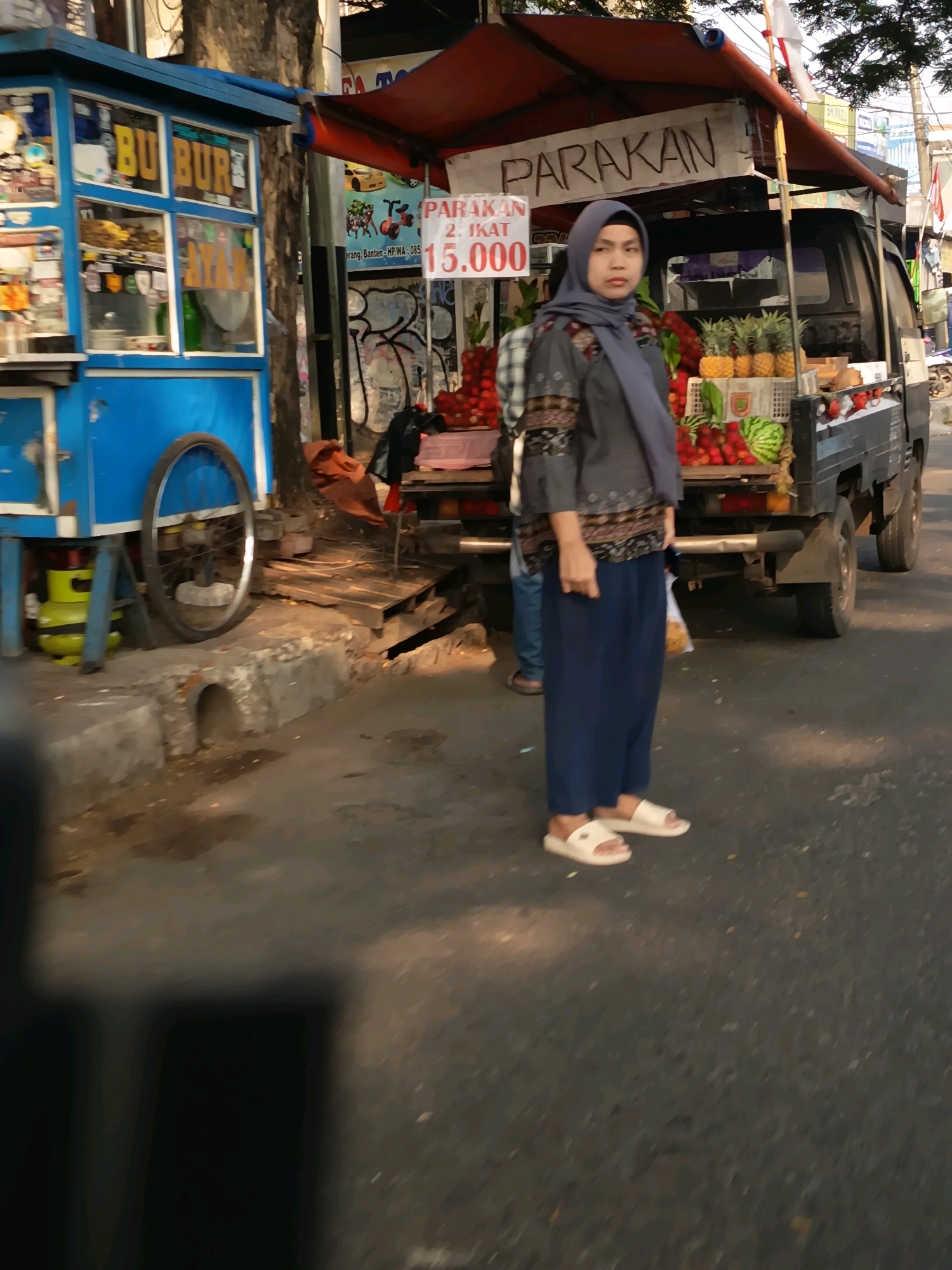 The image depicts a street scene in Tangerang, Indonesia, with a fruit stand and a food cart. A woman wearing a hijab and blue pants stands near the fruit stand. The fruit stand is loaded with various fruits, including pineapples and watermelons. A blue food cart with the words "BU BUR" and "AYAM" is visible on the left side of the image. A truck is parked behind the fruit stand, and the scene is set along a roadside with a paved surface and a sidewalk. The overall impression is of a bustling street market.