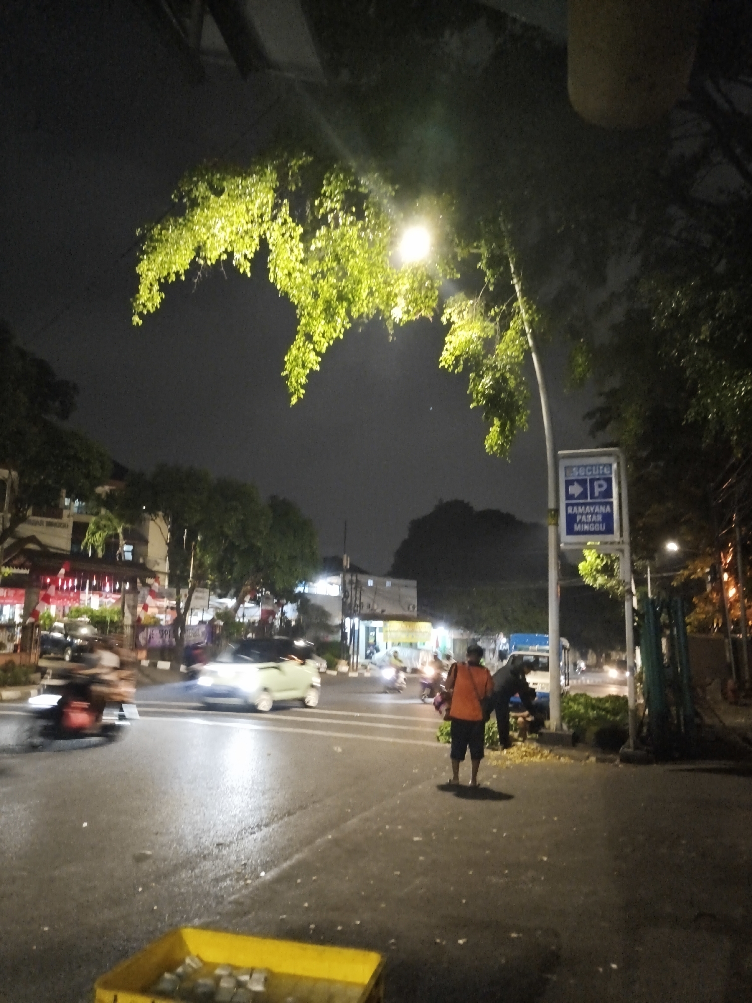 The image depicts a street scene at night in South Jakarta, Indonesia, likely near the Ramayana Pasar Minggu. A street lamp illuminates a lush green tree overhanging the road. In the foreground, a yellow crate is filled with white objects, possibly packaged goods. The wet asphalt suggests recent rain.
In the mid-ground, two individuals are present. One person, wearing an orange vest and dark shorts, stands facing away from the camera. Another person, also wearing dark clothing, is bent down near the base of a pole with a blue and white parking sign. This sign indicates "Secure" and provides directions for parking at "RAMAYANA PASAR MINGGU."
Several vehicles are on the street. A light-colored car is in motion on the left side of the road, its headlights on. To its left, a motorcycle with its headlight illuminated is also moving. Further down the street, other vehicles and lights are visible, indicating traffic. Buildings with illuminated windows and awnings are present on the left side of the road, suggesting commercial establishments. Trees and foliage line the street and sidewalk, with some leaves scattered on the ground. The overall atmosphere is that of a busy urban environment at night.