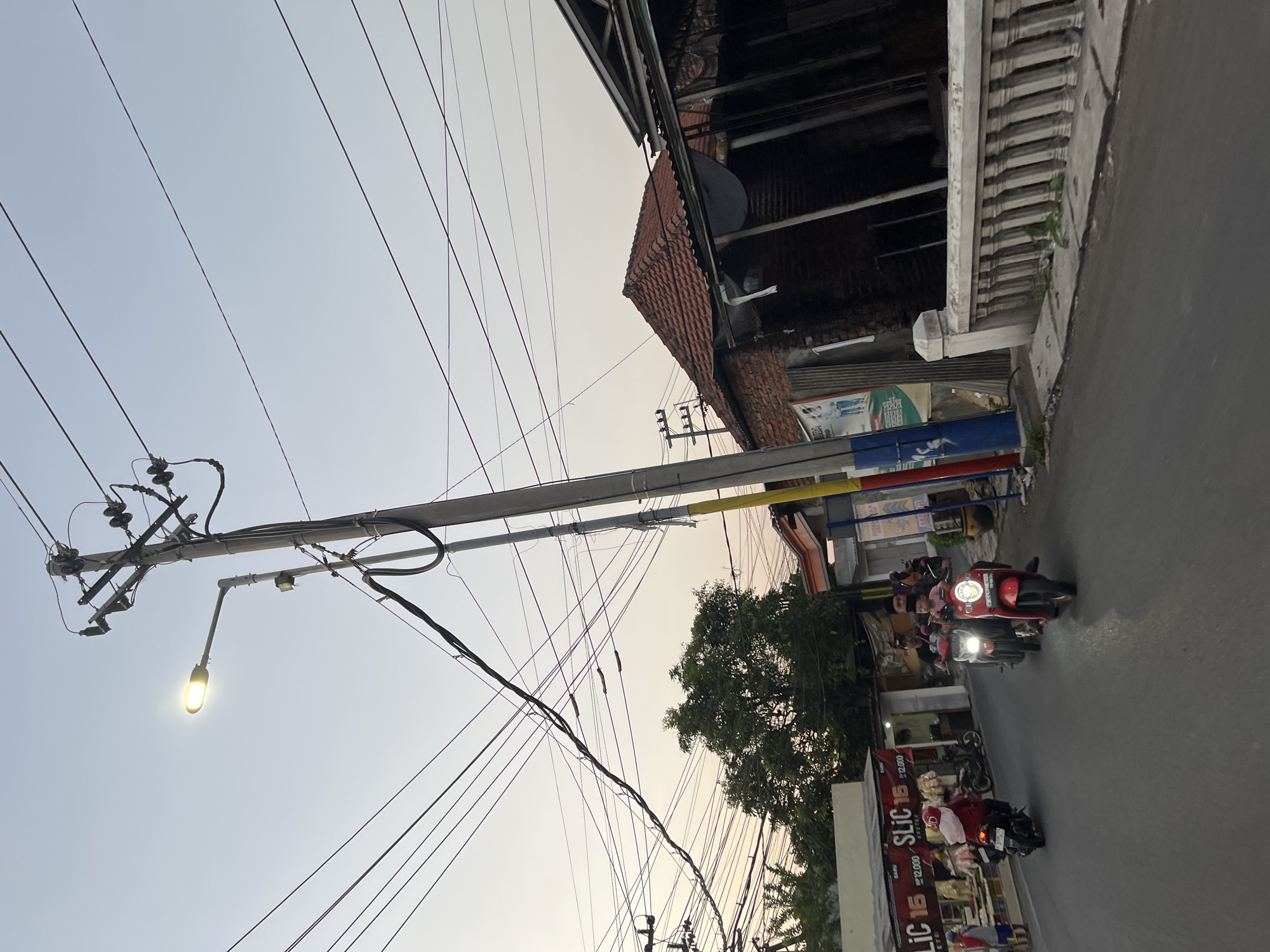 The image depicts a street scene in Karangtanjung, Indonesia, likely during the late afternoon or early evening, as indicated by the warm, soft light. The sky is a pale gradient of blue and orange, suggesting sunset or sunrise. The perspective is angled, with a prominent utility pole and a tangle of electrical wires dominating the left side of the frame. A street lamp on the pole is illuminated, casting a bright glow.
On the right side of the image, a street runs alongside buildings. The architecture appears to be a mix of traditional and modern, with a tiled roof on one structure and a more contemporary facade on another. There are several motorbikes on the road, with some people riding them, indicating a busy street. One red motorbike in the foreground is clearly visible. The overall atmosphere is that of an active urban environment transitioning into dusk.
Several signs with text are partially visible. One sign, partially obscured by a tree, appears to be colorful and possibly advertising. Another sign, more prominent on the lower right, has bold text that reads "SLIC 1B" and some smaller text that includes prices like "12,000". The presence of these signs, combined with the motorbikes and the general street activity, points to a commercial area. The weather appears calm, with no visible rain or strong winds. The image captures a slice of everyday life in this Indonesian locale.