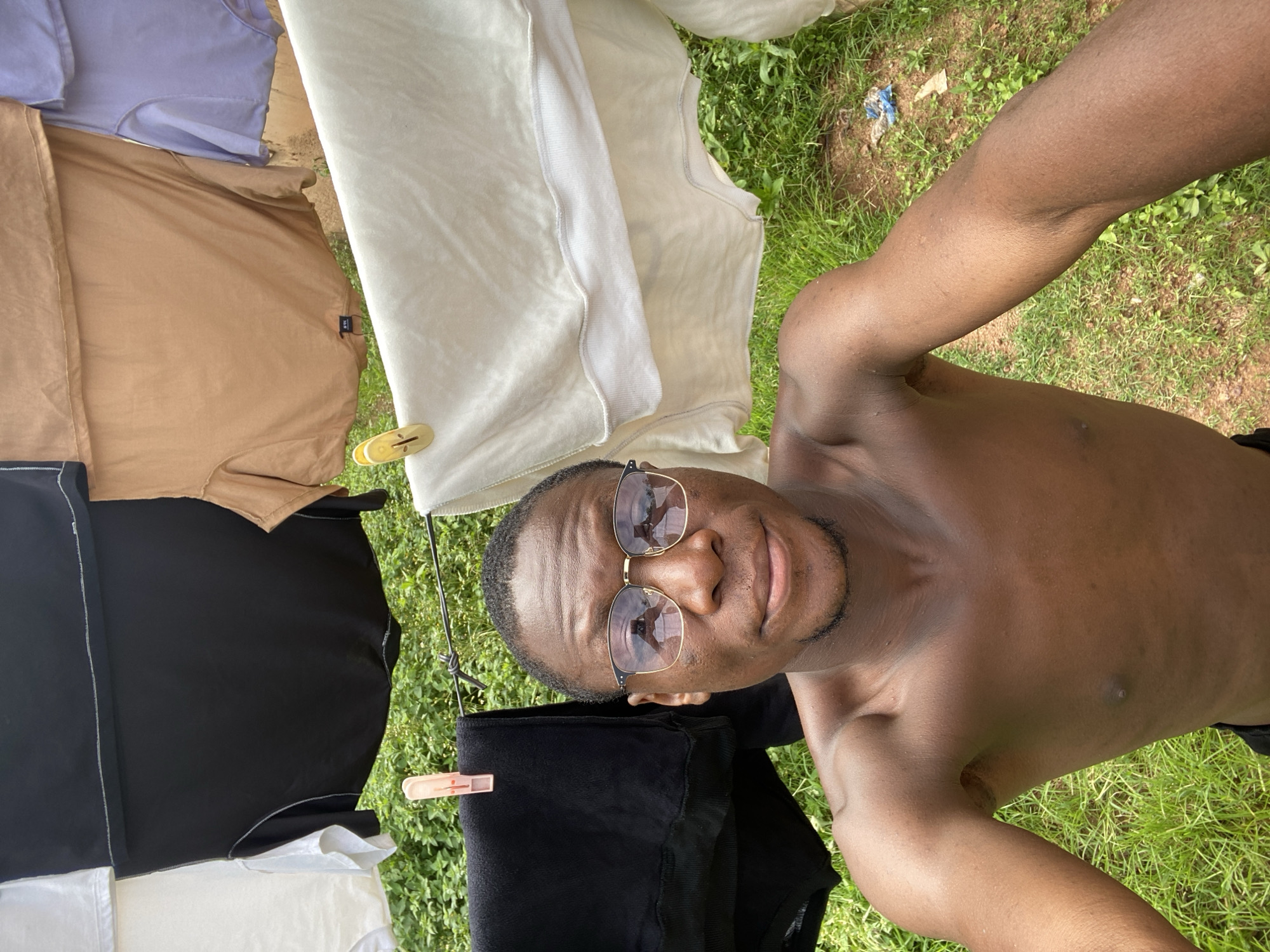 A young Black male wearing glasses and no shirt is taking a selfie outdoors in Bukuru, Nigeria. He is standing in front of a clothesline with several t-shirts hanging on it, including black, white, tan, and lavender shirts. The background is lush green grass and foliage. The lighting suggests it is daytime.