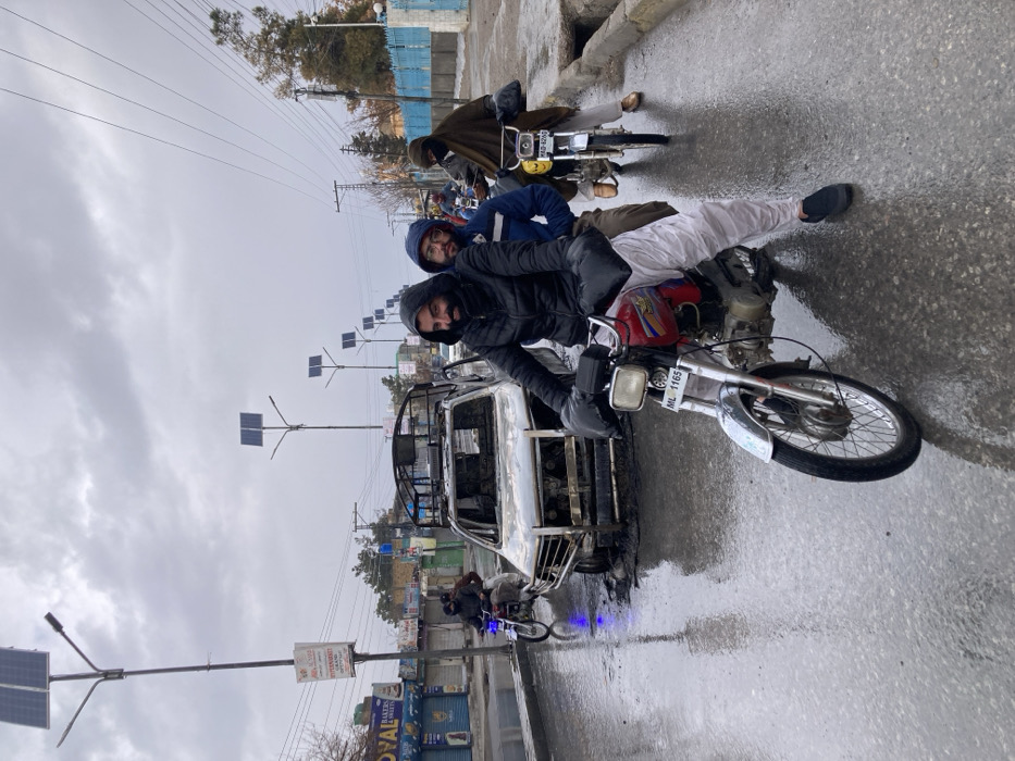 Three men ride motorcycles on a wet, grey day. A burnt-out vehicle sits at the edge of the road. A man in a black jacket drives the closest motorcycle with two passengers in the back, one in a blue jacket and another in white traditional clothing. The license plate reads "MIL 1165". Further down the road, another man drives alone on a separate motorcycle, wearing a dark brown robe. Streetlights with solar panels line the road.