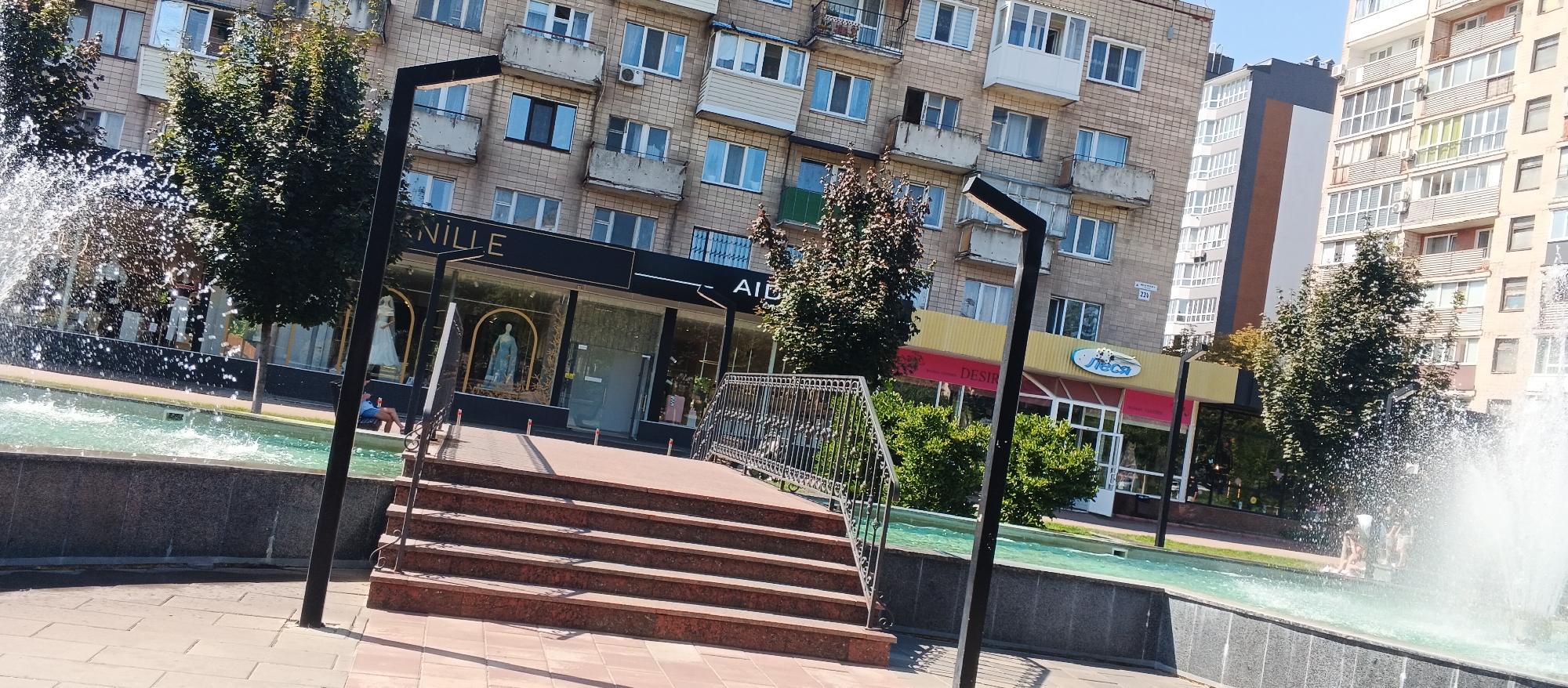 The image depicts a street scene in Cherkasy, Ukraine, with a focus on a modern building facade featuring shops and apartments. In the foreground, a fountain with splashing water creates a lively atmosphere. A set of stone steps leads up to a paved walkway. The shops have large glass windows displaying mannequins, suggesting clothing or bridal boutiques. One shop has a yellow awning with the Cyrillic text "ЛECA" and an oval logo. Another shop's sign reads "AINVILLE". Tall apartment buildings with balconies and multiple windows dominate the background. Trees with green foliage are interspersed throughout the scene. Several people are visible, including one sitting by the fountain and others inside or near the shops. The weather appears sunny and clear. The overall impression is an urban environment with commercial and residential elements.