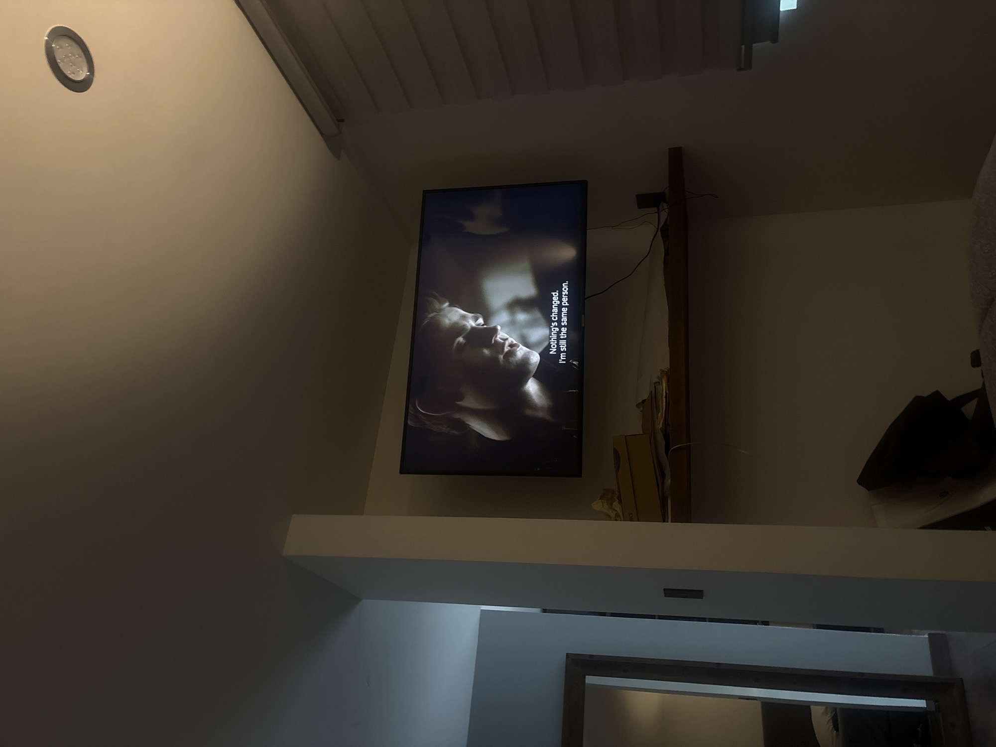The image shows a television mounted on a wall in an indoor setting. The television displays a scene with a person's face visible and text that reads, "Nothing's changed. I'm still the same person." The television is positioned above a white shelf or ledge. Above the television is a ceiling with a slatted design. The setting appears to be a home or living space. The image does not contain enough information to locate it precisely in San Bernardino Tlaxcalancingo, Mexico.