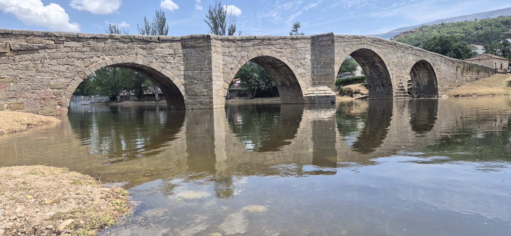 This is a landscape photograph of a stone bridge spanning a body of water under a partly cloudy sky. The bridge is constructed from large, rough-hewn stones and features several arches. The water in the foreground is calm, with reflections of the bridge and sky visible on its surface. The banks of the water are lined with sand and some vegetation. In the background, there are trees, some buildings, and rolling hills covered in greenery. The overall scene depicts a serene, natural setting.