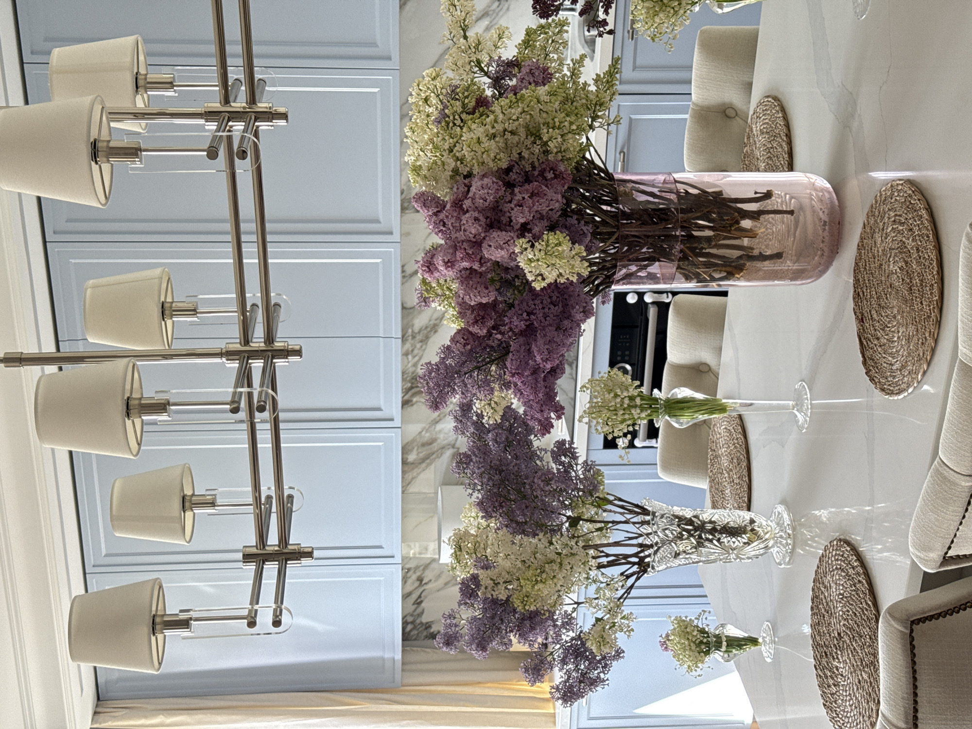 A chandelier hangs above a dining table set with round placemats, vases of flowers, and chairs. The room has blue cabinets with white trim. The flowers are predominantly purple and white lilacs. The background may be a kitchen or dining room. The location is likely Moscow, Russia.