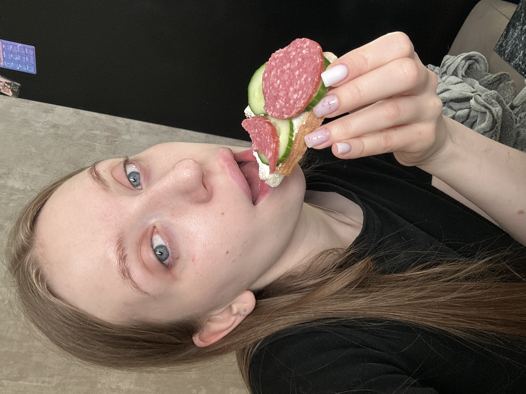 A young woman is lying down while holding a sandwich towards her mouth. The sandwich consists of bread, white spread, cucumber slices, and salami. She has light skin, blue eyes, and light brown hair. She is wearing a black shirt. The background includes a textured gray surface, a black wall, and a gray, rumpled fabric or blanket. A blue object and other items are visible on the gray surface. The city of Chaikovsky, Russia is not identifiable in this image.