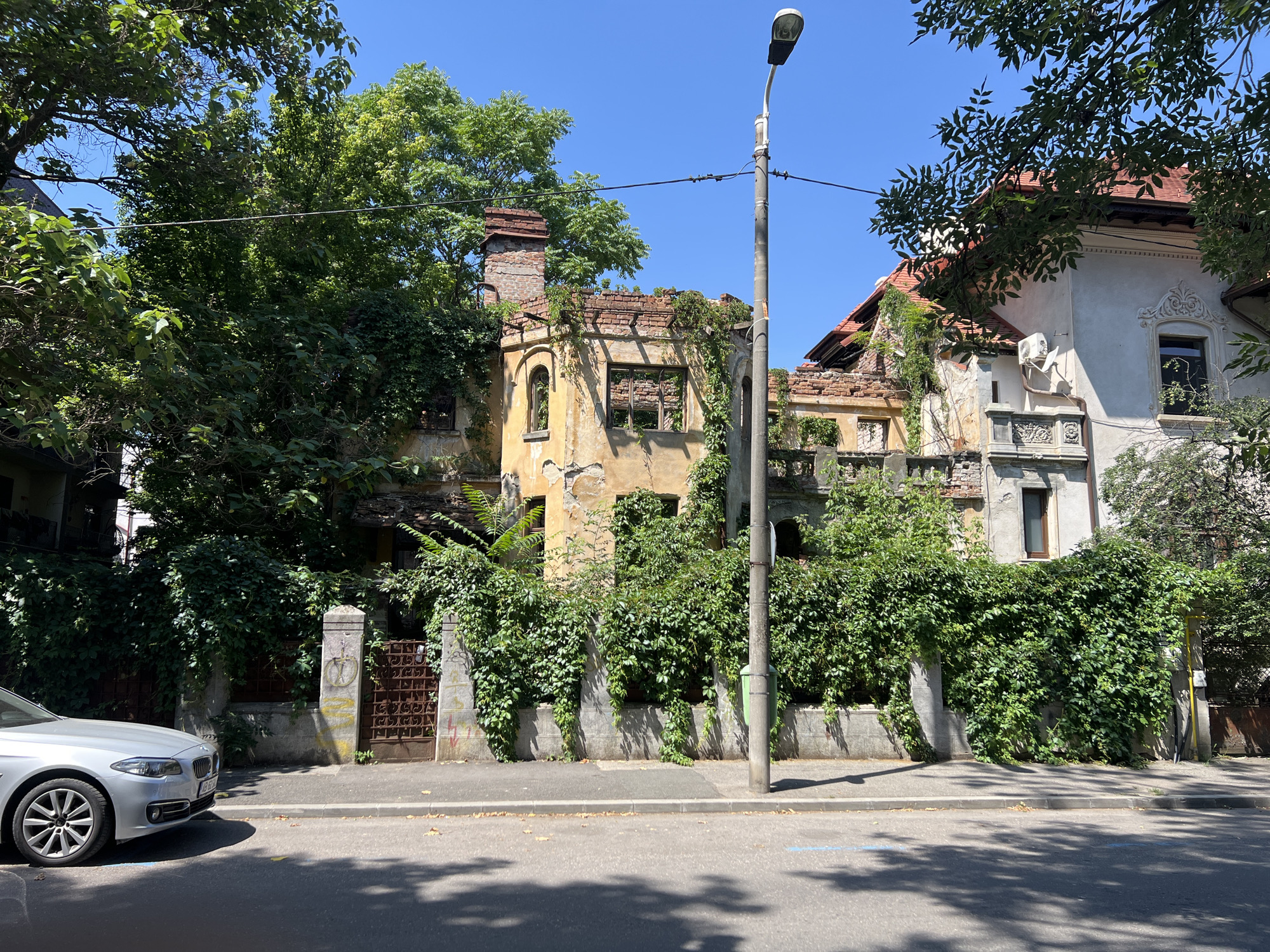 In Bucharest, Romania, a silver sedan is parked on the side of a street in front of an old, dilapidated building. The building is partially overgrown with ivy and appears to be abandoned. The architecture suggests it may be a historic structure. A tall lamppost stands on the sidewalk in front of the building. The sky is clear and blue, indicating a sunny day. The street is paved, and there are shadows cast by trees and possibly the building onto the road.