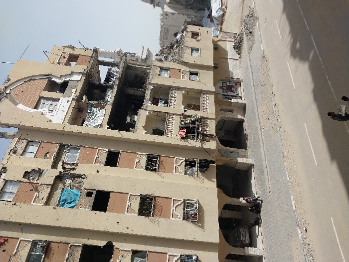 The image presents a street scene in Khan Yunis, Palestinian Territory, dominated by a heavily damaged multi-story building. The building, a light tan structure with dark brown accents, shows extensive destruction across its facade, including large missing wall sections that expose interior rooms on multiple floors. Numerous windows are broken or entirely absent, and the exterior surface is pockmarked with apparent bullet or shrapnel impacts. Balconies exhibit damage, with some containing debris or hanging fabric, including a prominent blue tarp on a middle-left balcony. An arched decorative element on the upper right side of the building is also significantly compromised.
At ground level, a paved road with visible white double-line markings extends across the foreground. A light-colored paved sidewalk borders the building. Beneath the building's arched ground-floor supports on the left, a silver sedan is parked. Three individuals are on the sidewalk near the vehicle; two appear to be crouched or seated, while one stands nearby. Further to the right along the road, piles of rubble and construction debris are present. In the immediate foreground, two additional individuals are walking away from the camera, one carrying a white package. The background on the right reveals further damaged structures, several white tents, and the partial view of a white dome structure, possibly a mosque, under a hazy sky.