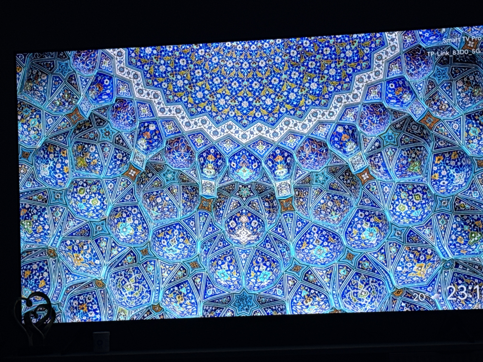 The image is a close-up of a TV screen displaying a complex, symmetrical Islamic geometric pattern. The pattern is primarily composed of blue, white, and yellow floral and star-shaped motifs. The background color is a rich blue, with the other colors highlighting the intricate details of the design. The style and patterns suggest it might be inspired by the Shah Mosque in Isfahan, Iran.
On the top right corner of the screen, text displays "Smart TV Pro TP-Link_83DO_5G". In the bottom right corner, the screen shows a digital display with "20°" and "23:11" indicating temperature and time.
In the lower left corner of the picture, outside of the screen, a dark object shaped like intertwined hearts is visible. To its right, a small white object can also be seen. The scene is dimly lit.