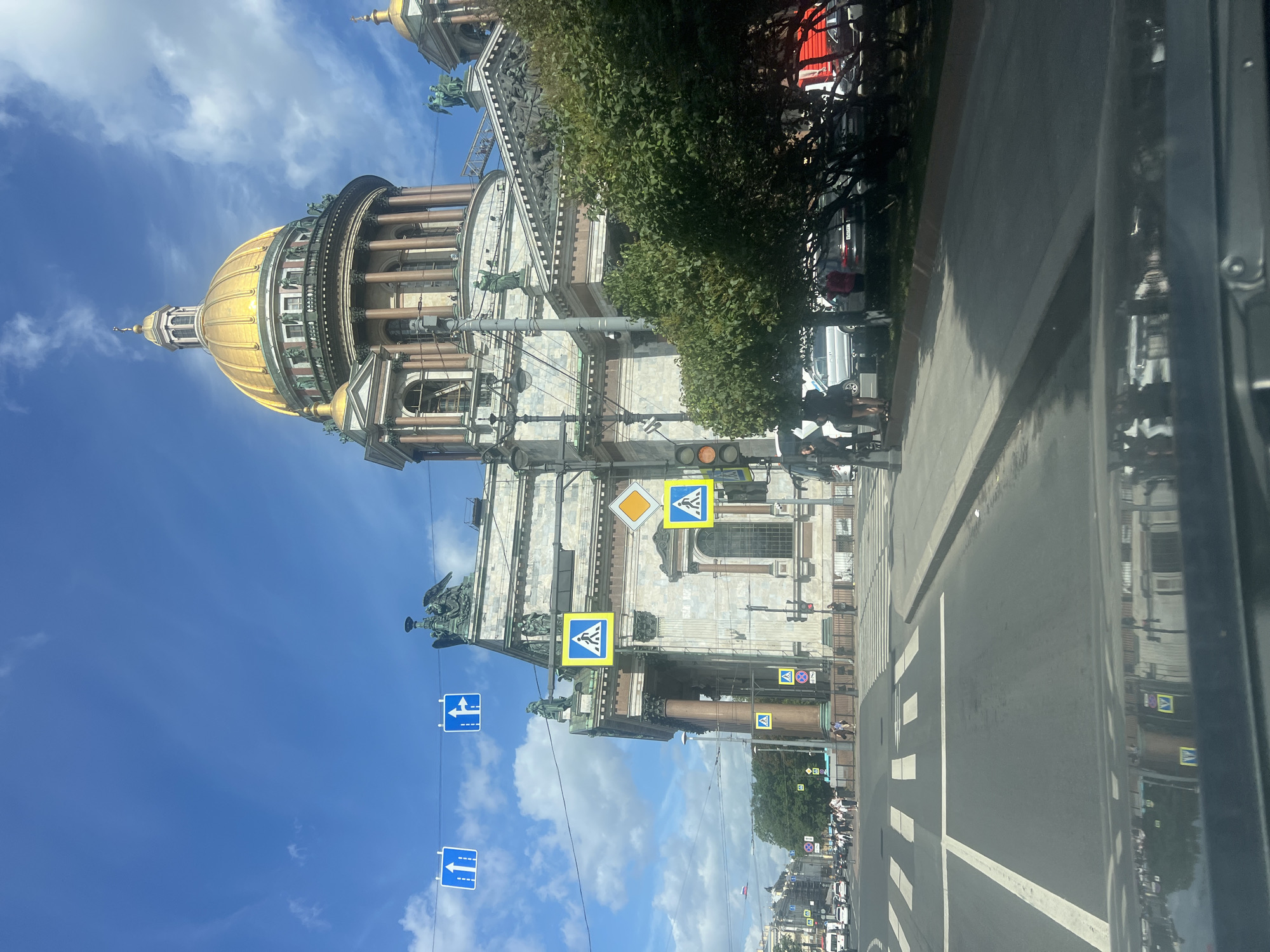 The image shows a view from inside a vehicle looking out at St. Isaac's Cathedral in Saint Petersburg, Russia, on a bright, partly cloudy day. The grand cathedral, with its golden dome, is a prominent feature, partially obscured by trees and scaffolding on its facade. In the foreground, a street with crosswalks and traffic lights is visible, along with cars and pedestrians. Several blue traffic signs, indicating lane direction and pedestrian crossings, are suspended over the road. The reflection of the cathedral and surrounding buildings is visible on the vehicle's dashboard. The overall scene depicts a typical urban environment with historical architecture and everyday activity.