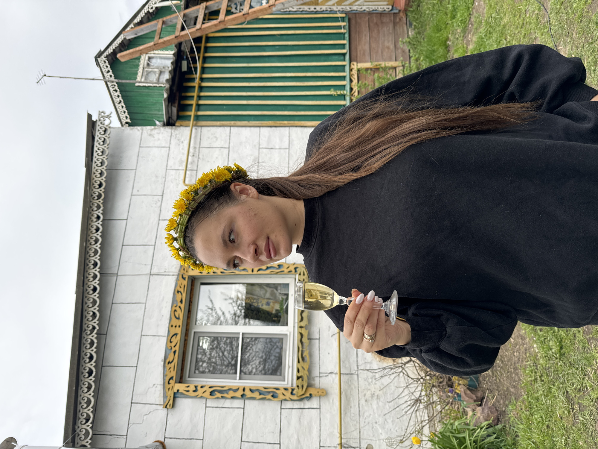 A woman wearing a dandelion wreath on her head holds a glass of champagne. She is wearing a black sweatshirt. She is standing in front of a building with a green and white facade. The building is located in Красный Маяк, Russia. There is grass and a few flowers growing in front of the building. The sky is overcast.