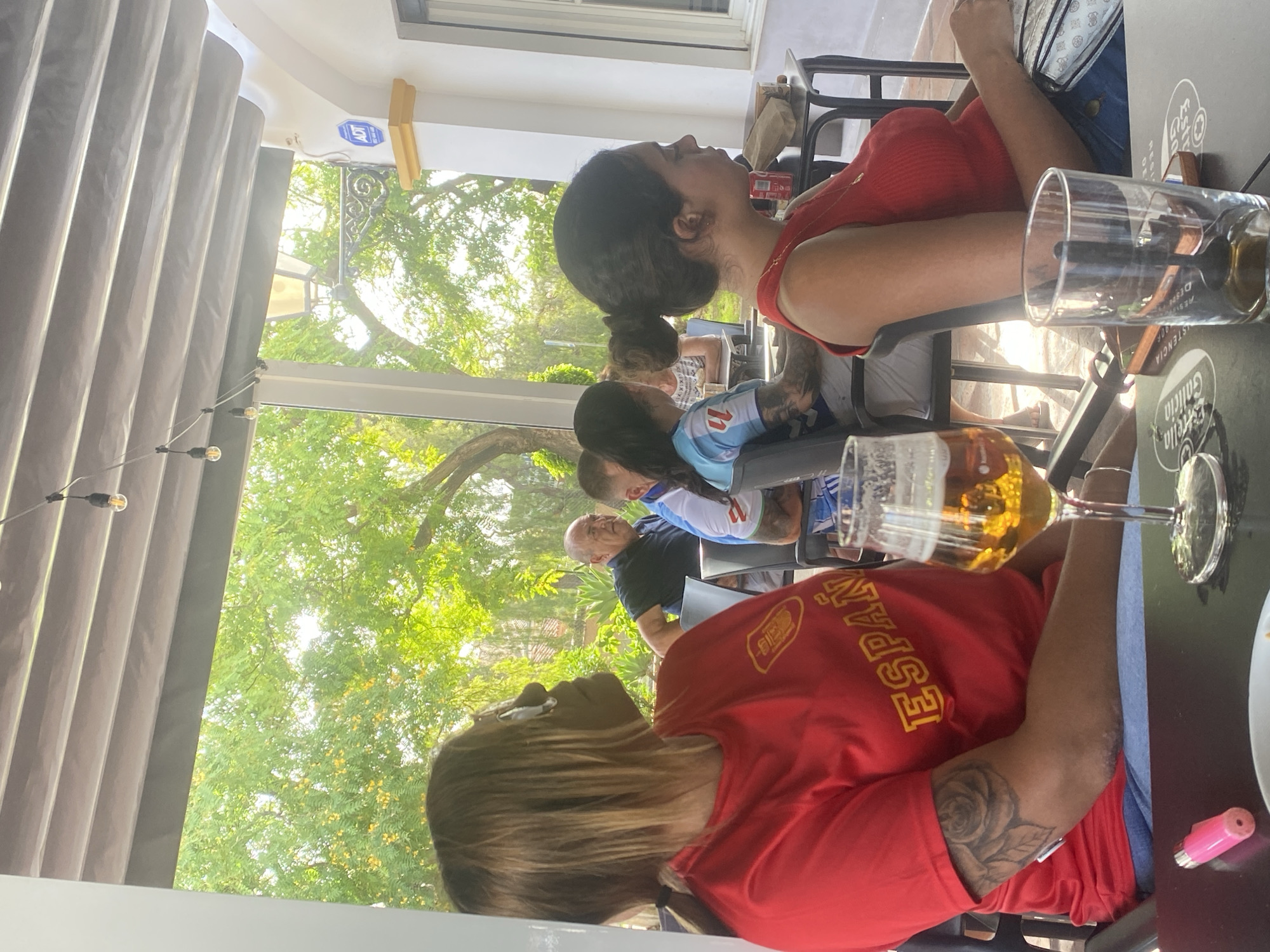 In San Pedro De Alcántara, Spain, an outdoor seating area of a restaurant is captured. The scene features several people seated at tables, some appearing to be engaged in conversation or watching something. In the foreground, a woman wearing a red shirt with the word "España" prominently displayed is seen from the chest up, with a tattoo on her arm. To her right, another woman in a red top is seated, with her hair styled in a bun. A glass of amber-colored liquid, likely beer, is placed on the table in front of the first woman. In the background, other patrons are visible, including a man in a dark shirt and a person wearing a blue and white sports jersey. The environment is characterized by a shaded patio with trees and greenery visible in the background, suggesting a sunny day. An ADT security sign is mounted on a white pillar to the right of the image.