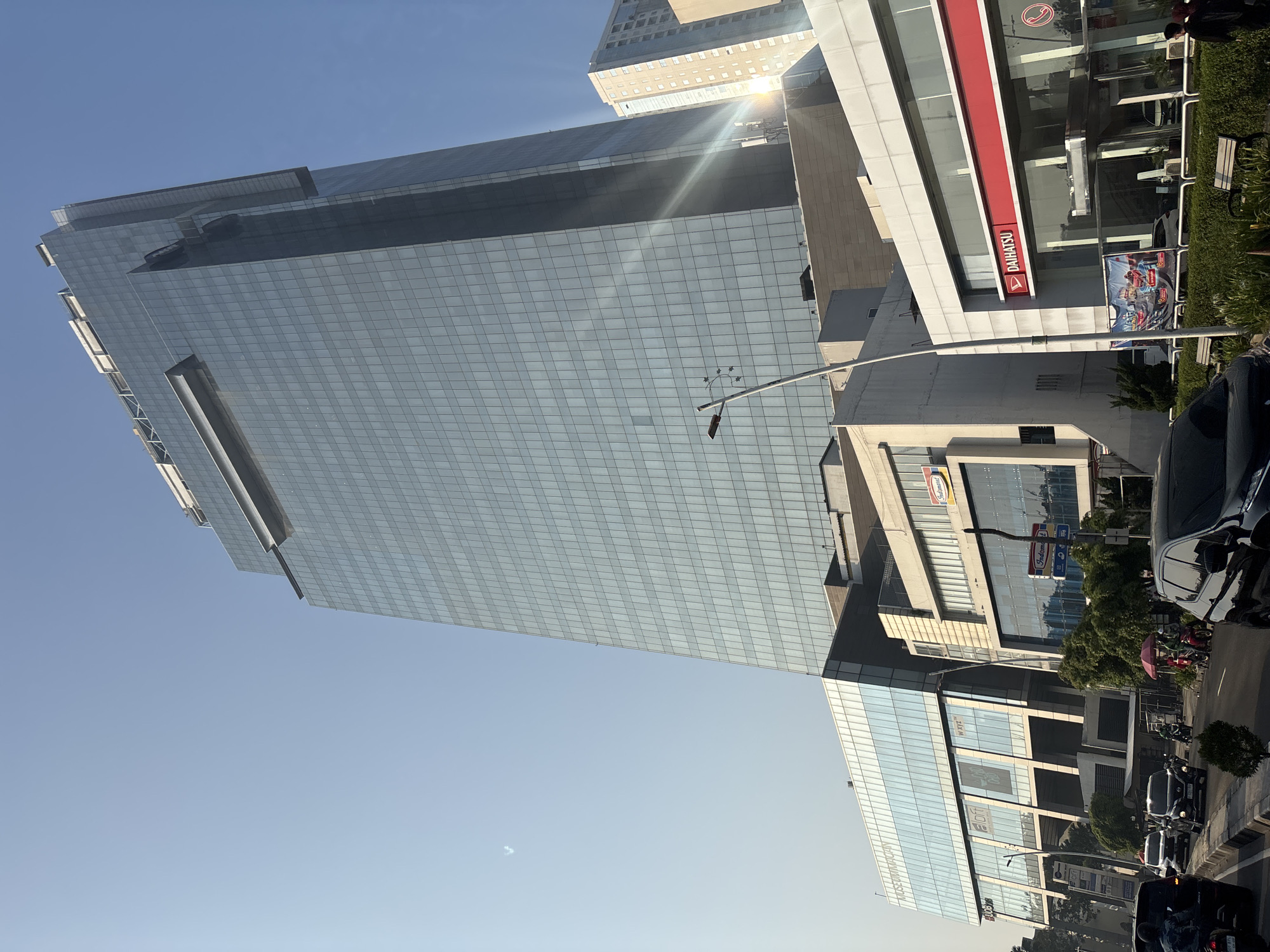 The image shows a street view in West Jakarta, Indonesia, with a prominent glass-covered skyscraper dominating the background. The skyscraper has a sleek, modern design with a tiered structure. In the foreground, a busy street is visible with several cars and motorcycles driving. Several buildings line the street, including what appears to be a retail establishment with a red and white "Daihatsu" sign. The sky is clear and blue, with the sun casting a bright glare on the skyscraper. There are trees and some outdoor seating areas visible along the sidewalk.