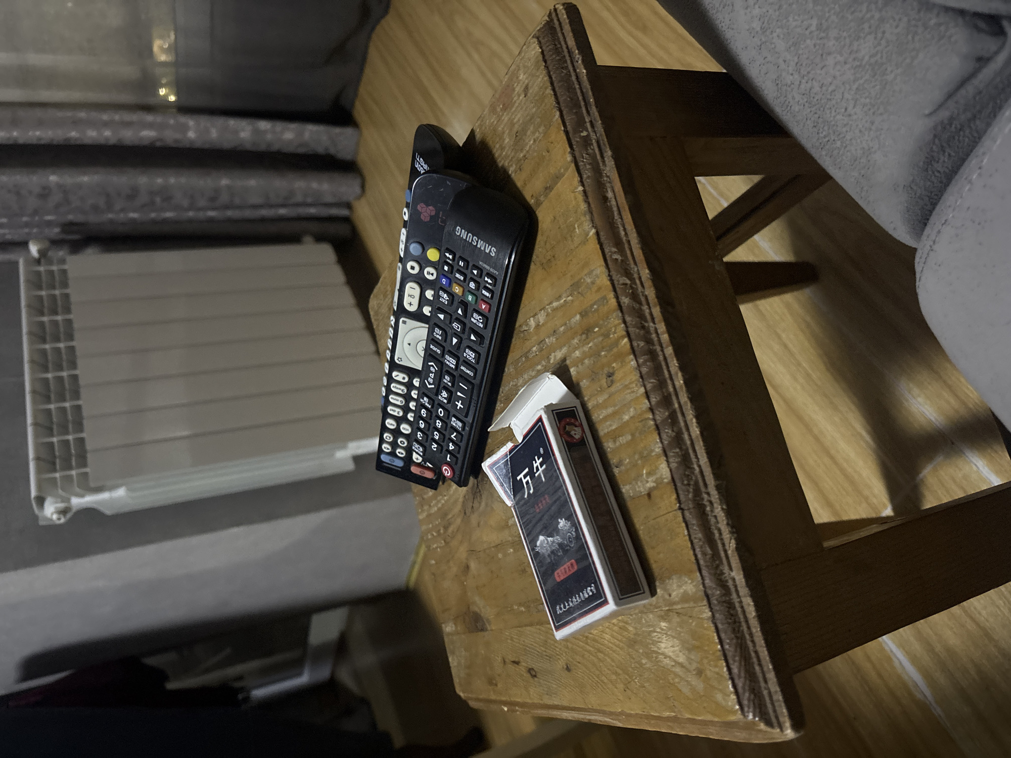 A Samsung remote, a pack of "Wan Niu" cigarettes, and a white, rectangular object sit on a wooden surface. The lighting in the scene is dark.