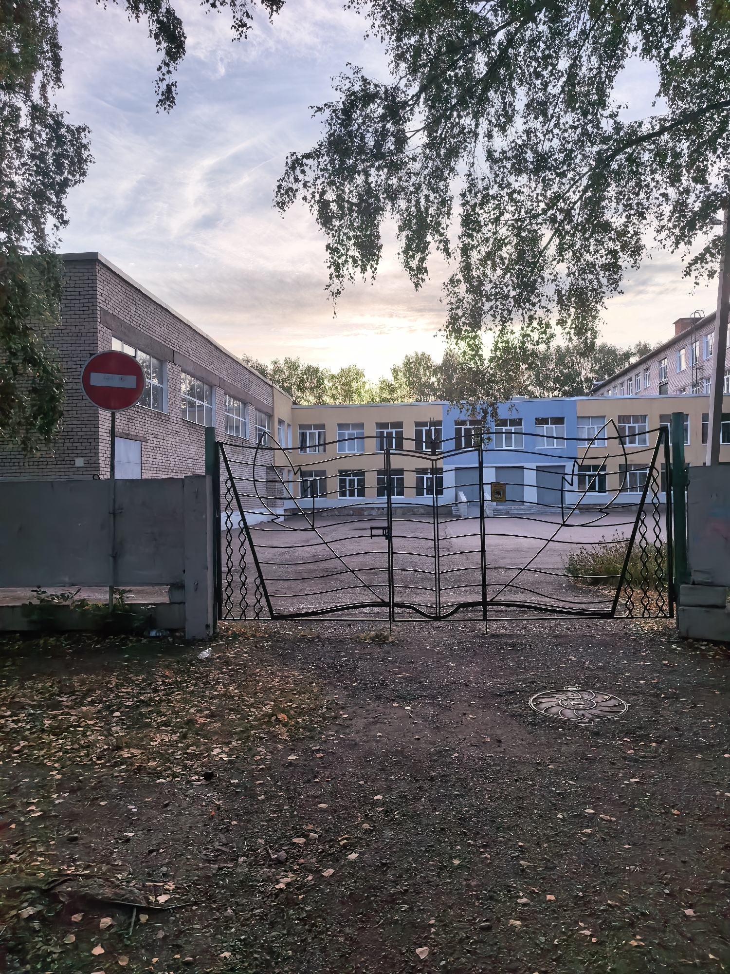 The image depicts the entrance to a school complex in Sterlitamak, Russia. The foreground is a dirt path covered with fallen leaves, indicating an autumnal setting. A dark metal gate with a decorative, wavy pattern stands centrally, partially open, leading into the school grounds. To the left of the gate is a grey concrete fence and a red circular road sign with a white horizontal bar, signifying "no entry." Behind the gate, a school building is visible, characterized by its multi-colored facade – sections of yellow, light blue, and grey. Multiple windows with white frames are apparent on the building. Trees with green foliage are present on the left and right sides of the image, with some branches hanging over the scene. The sky above is partly cloudy, with streaks of white clouds against a pale blue backdrop, suggesting it is either early morning or late afternoon. The overall impression is a quiet, possibly deserted school entrance.