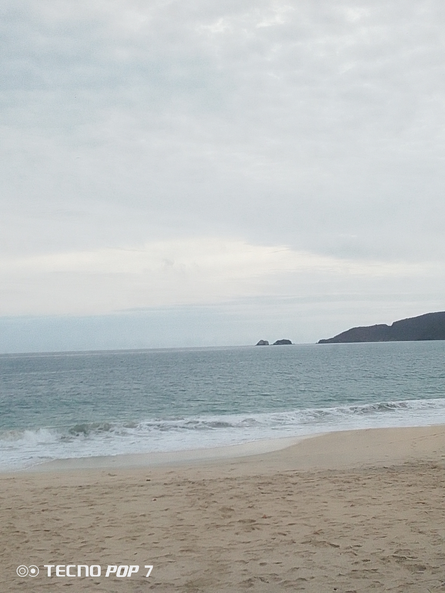 The image shows a beach in Pedro González, Venezuela on an overcast day. The scene includes a sandy beach in the foreground, light-blue ocean waves breaking on the shore, and land with rocky outcroppings in the distance. The sky is a uniform gray. There are no people visible in the image. A watermark of the phone model, TECNO POP 7, is in the lower left corner.