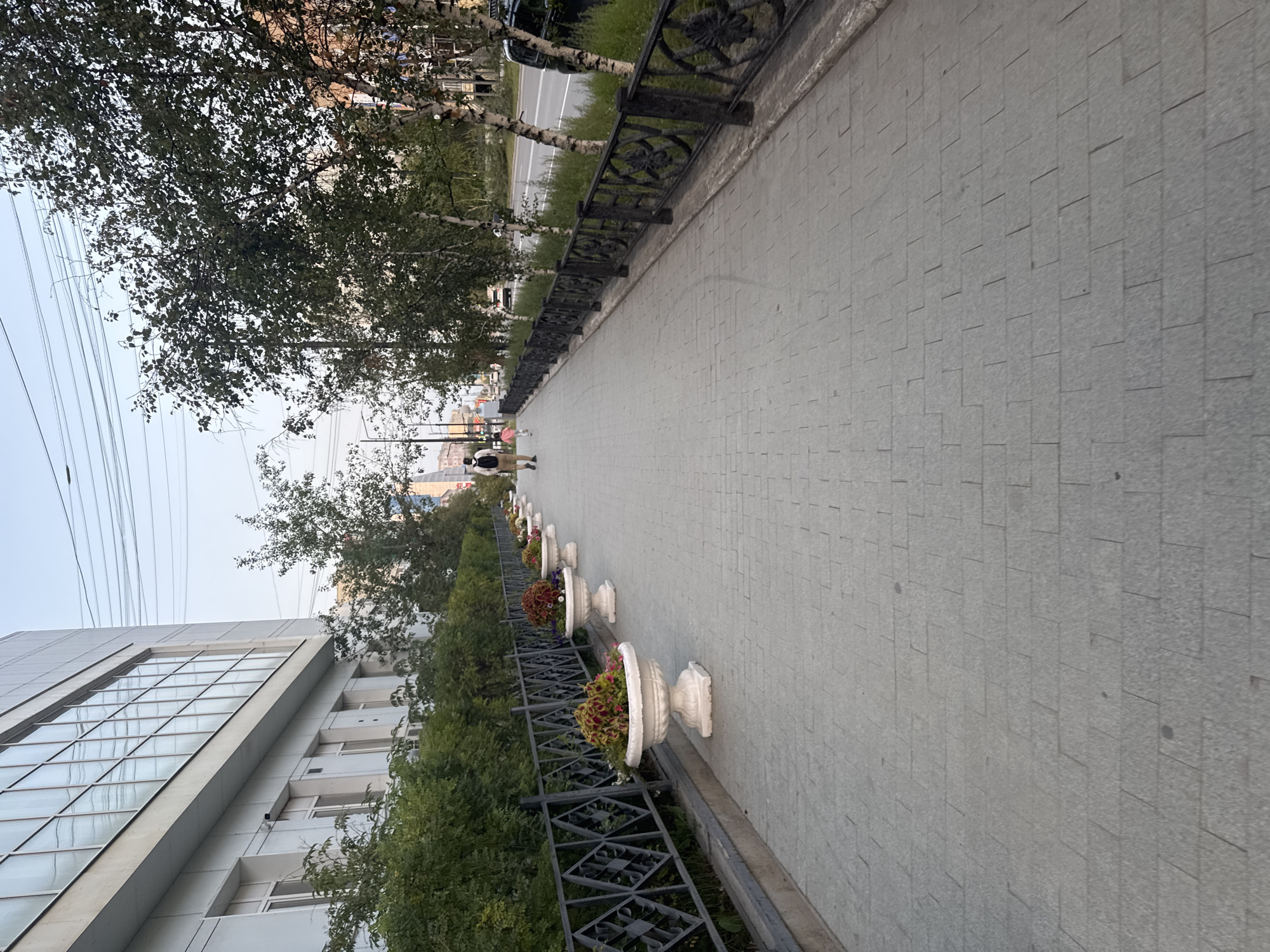 The image depicts a sidewalk in Yakutsk, Russia, with a modern building on the left and trees lining the street on the right. The sidewalk is paved with gray rectangular stones laid in a stretcher bond pattern. Along the sidewalk, a black metal fence separates it from a row of white, ornate planters filled with colorful flowers. Two individuals, a man carrying a backpack and a young child, are walking away from the camera on the sidewalk, further down towards the street. A road with some vehicles is visible beyond the trees. The sky is a pale blue, suggesting it is either early morning or late evening.