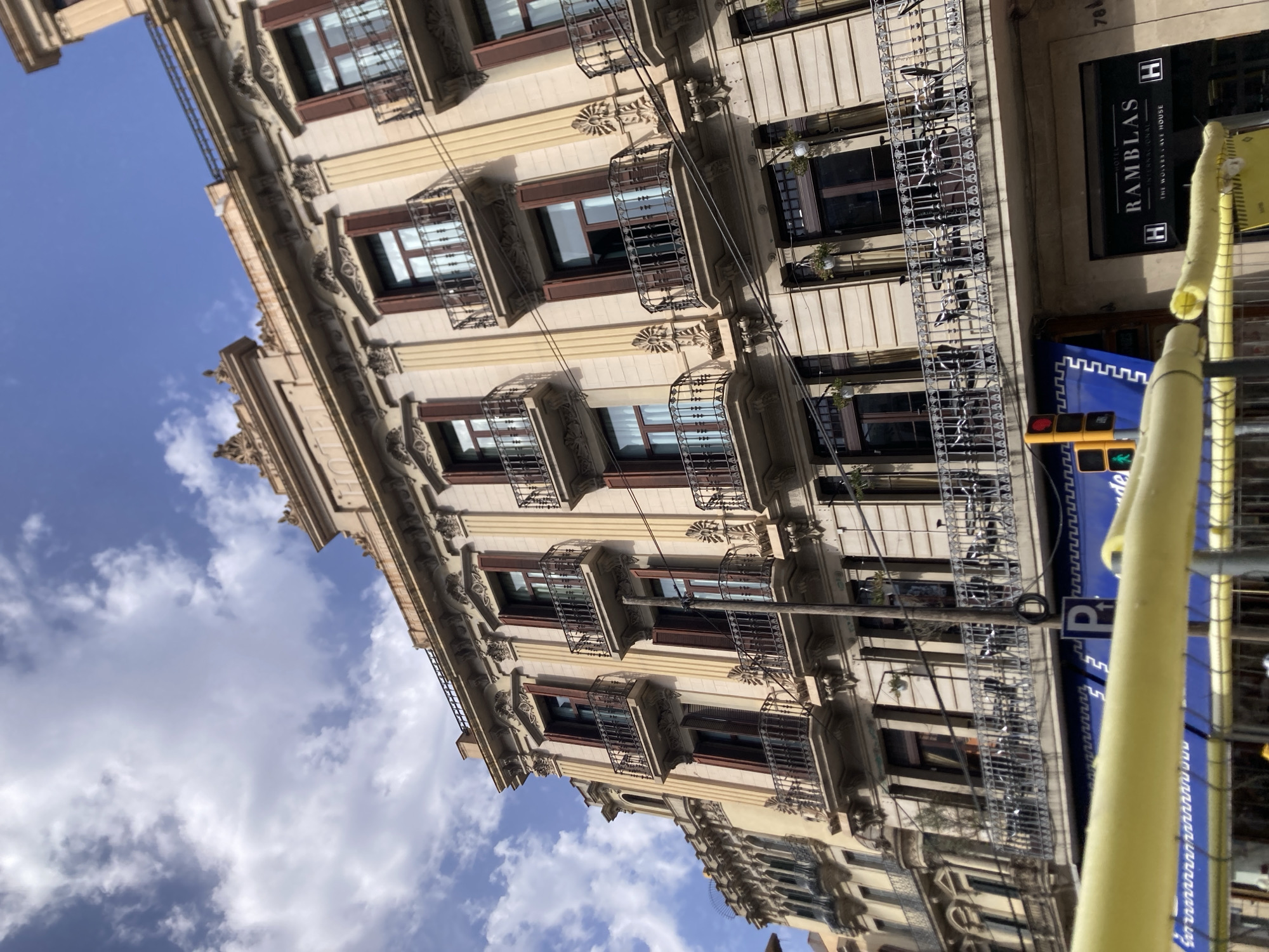 The image shows a close-up, low-angle view of a historic building facade in Barcelona, Spain. The building is ornate, with classical architectural details, including columns, decorative molding, and multiple balconies with wrought-iron railings. The facade is painted a pale yellow. The balconies are furnished with chairs and tables, suggesting a cafe or restaurant on that level. Above, the sky is blue with scattered white clouds.
Below the balconies, the ground floor features a blue awning with white lettering and a traffic light with red and yellow lights. A sign for "HOTEL RAMBLAS INTERNACIONAL" is visible to the right of the blue awning, along with the text "THE WOLVES CAVE HOUSE." The presence of "Ramblas" in the sign and the style of the architecture strongly indicate the location is on La Rambla, a famous boulevard in Barcelona. Yellow protective barriers are in the foreground, obscuring the very bottom of the image. The scene is captured on a clear day.