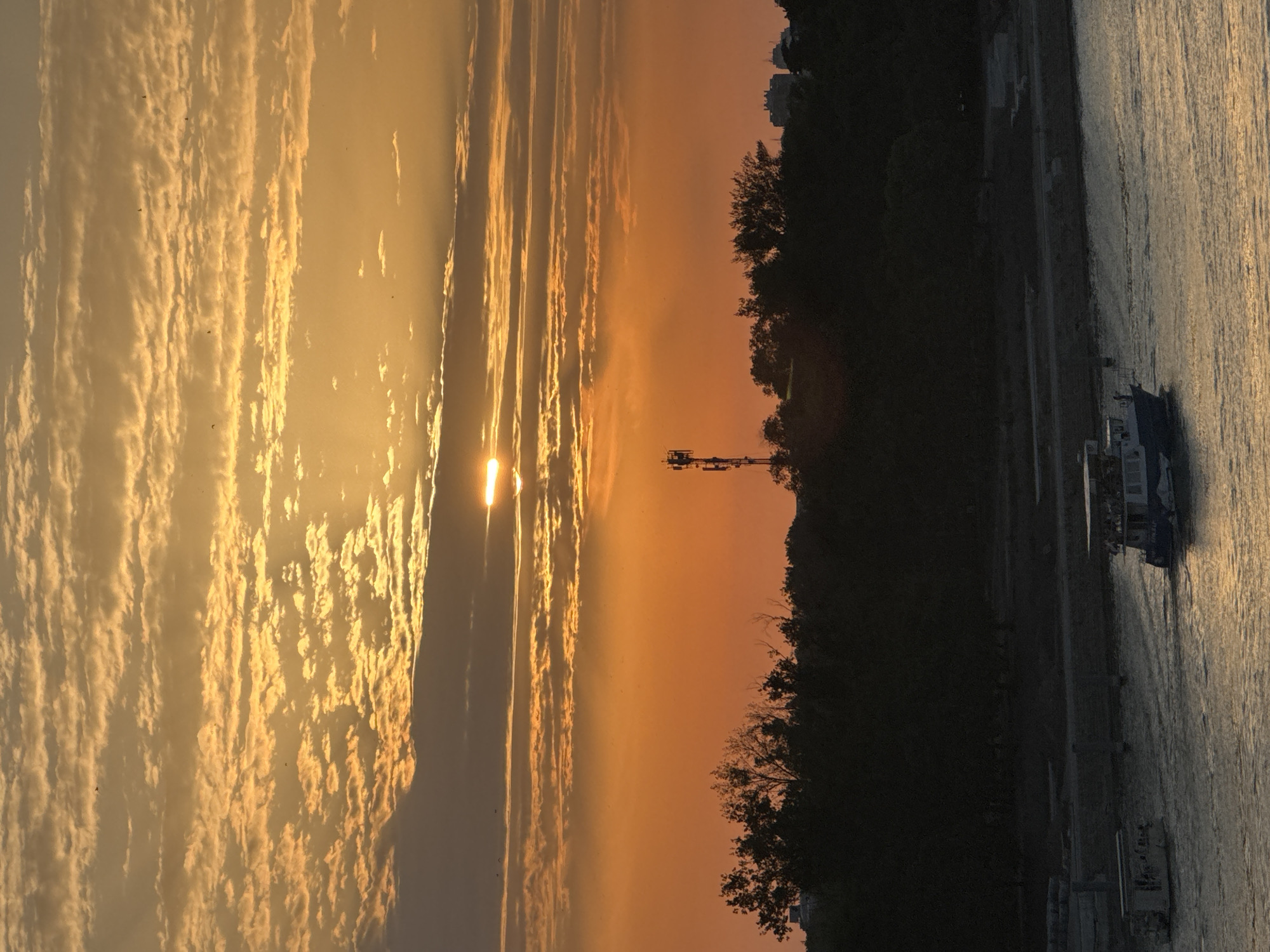 The image captures a sunset over the Sava River in Belgrade, Serbia. The sky is filled with dramatic orange and yellow clouds, with the sun peeking through them. In the foreground, the water of the Sava River reflects the warm hues of the sunset. Two boats are visible on the river. The boat on the right is larger and appears to be a tour boat, carrying several passengers on its deck. The boat on the left is smaller and less populated. Along the riverbank, a dark silhouette of trees and vegetation forms a dense line. A communication tower stands out against the sky behind the trees, and in the distance, the faint outlines of buildings can be seen. The overall scene conveys a peaceful and serene atmosphere as day transitions into night.