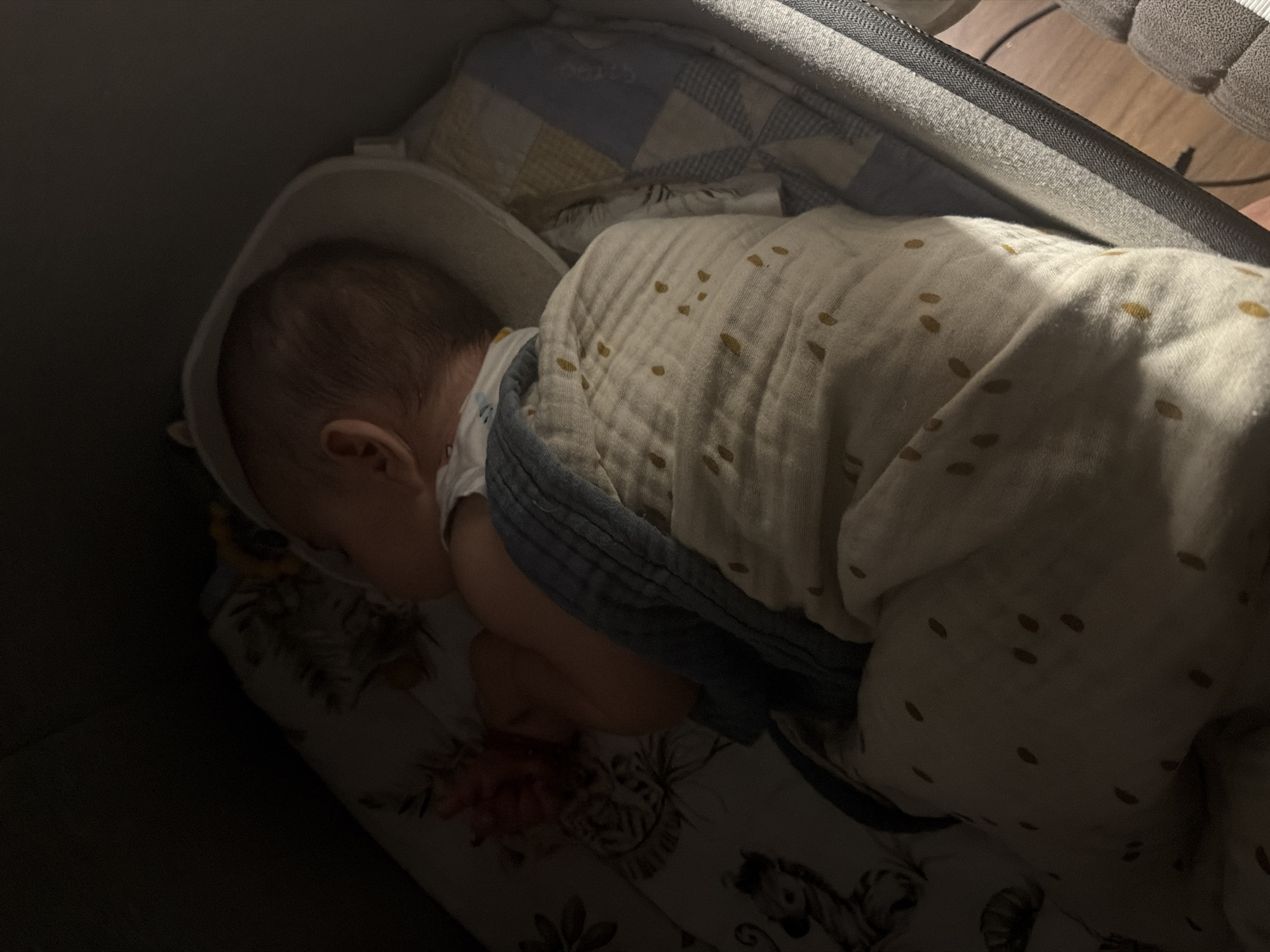 A newborn infant is sleeping in a bassinet in Pontonx-sur-l'Adour, France. The infant is swaddled in a light-colored muslin blanket with scattered mustard-yellow dots and is wearing a white onesie. A blue-grey textured blanket is partially visible under the muslin blanket. The bassinet has a padded rim and patterned sheets featuring animals and floral motifs on the left side, and a quilted pattern of blue and yellow squares on the right. The overall lighting is dim, with a light source illuminating the infant from the right side, casting shadows. The scene appears to be indoors, likely in a bedroom.
