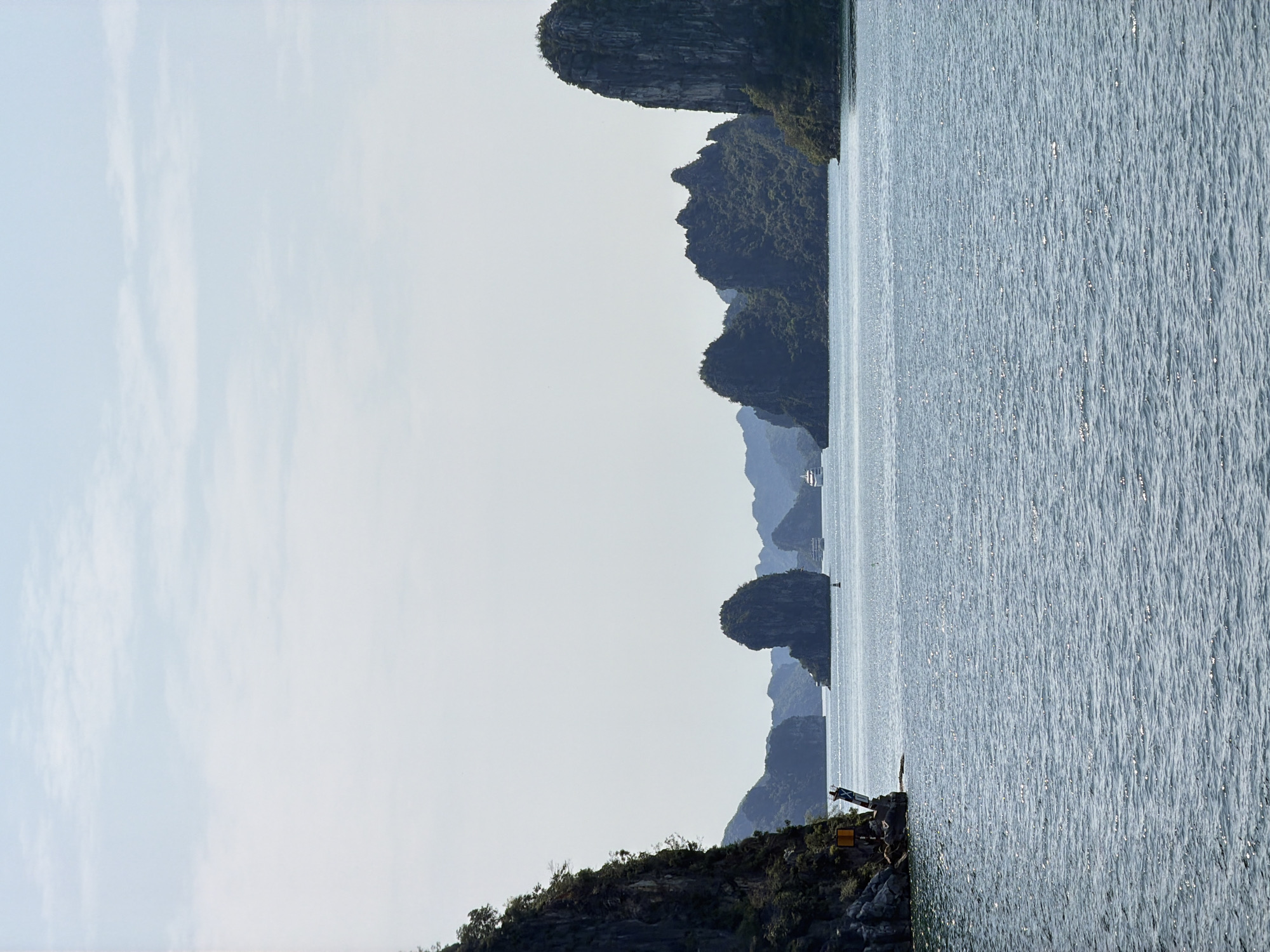 The image depicts a seascape with limestone karsts, likely in Ha Long Ward, Vietnam. The karsts are covered in green vegetation. A body of water occupies the right side of the frame, its surface reflecting sunlight. The sky is visible on the left side, with a light, overcast appearance. In the lower right corner, near the water's edge, a construction site or quarry is visible with heavy equipment.