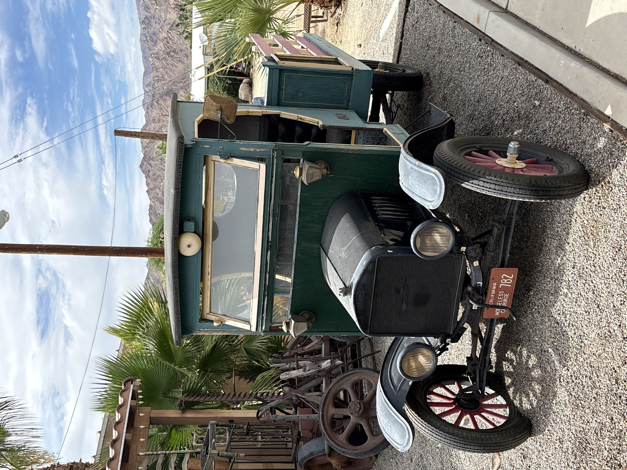 The image shows a vintage green and black truck parked on a gravel surface with a concrete curb to the right. The truck has a wooden bed with pink and red railings. It features large, spoked wheels with red hubs and a black grille with the Ford logo. A California license plate reading "HOMELESS CARRIAGE 782" is attached to the front bumper. In the background, there are palm trees, a rustic wooden structure with outdoor lighting, a wooden pole with wires, and mountains in the distance. The sky is cloudy. The scene appears to be in an outdoor setting, possibly a roadside attraction or a historical site. The location is Chiriaco Summit, United States.